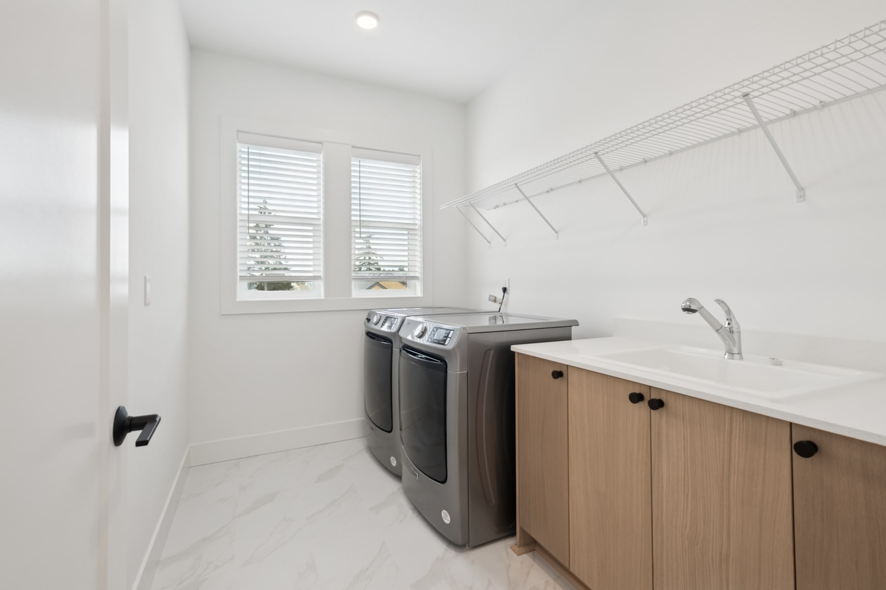 A modern and minimalist laundry room with white walls, a large window, and wooden cabinets housing a washer and dryer.