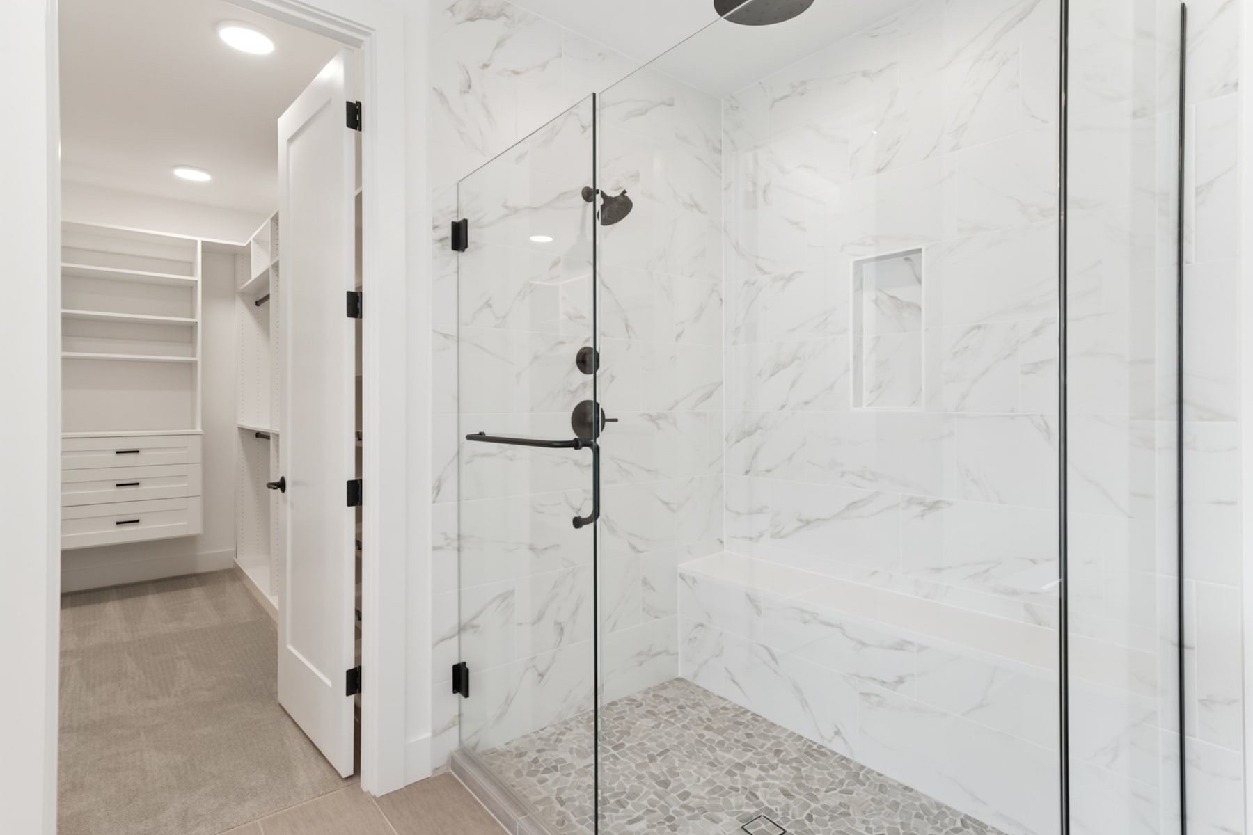A modern, minimalist bathroom with a glass shower enclosure, white marble walls, and a built-in closet in the background.