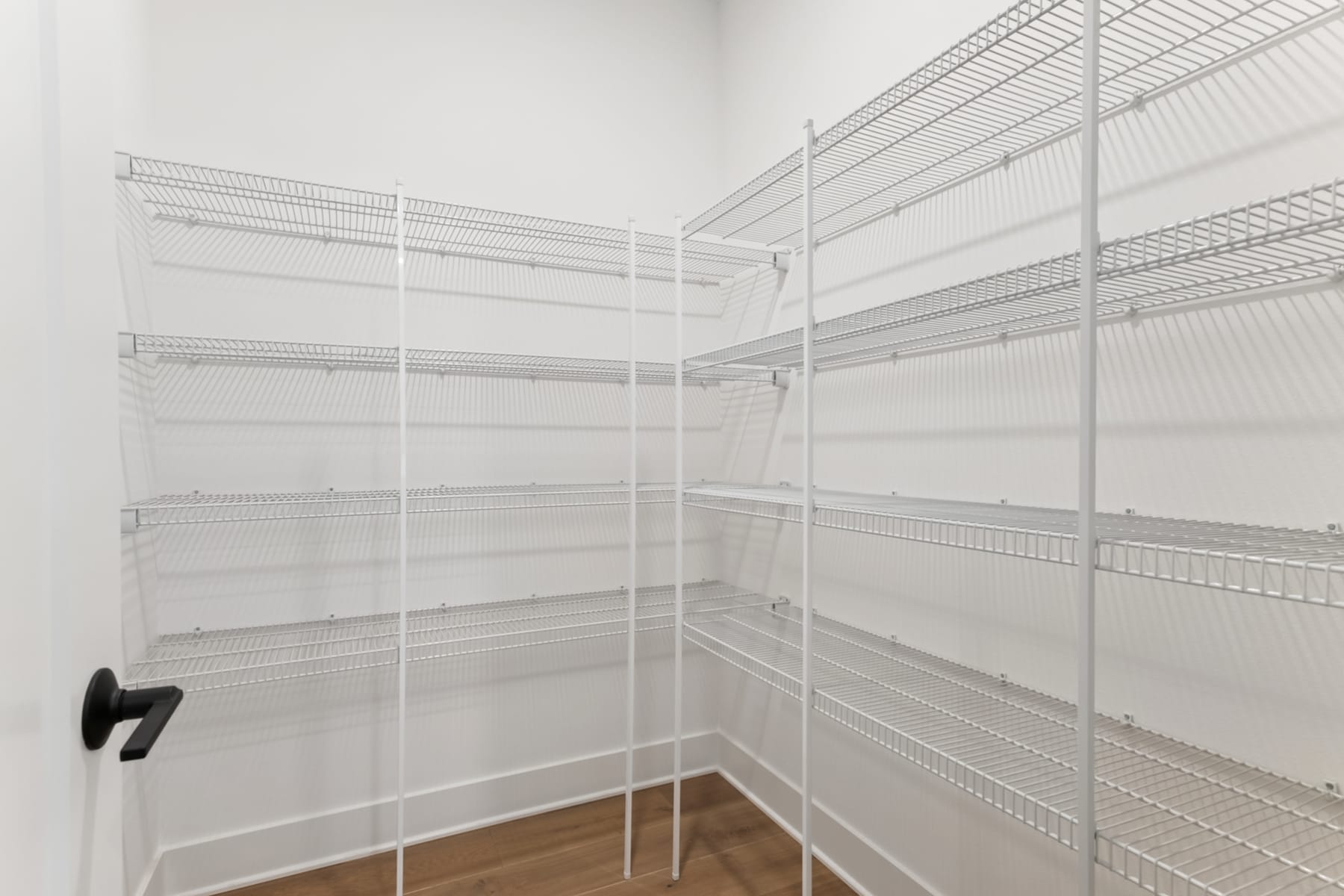 A white wire shelving unit with multiple levels, situated in a corner of a room with a wooden floor.