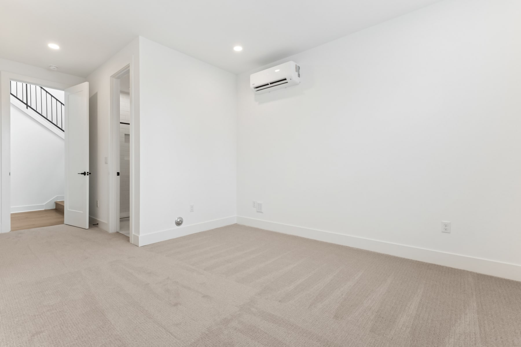A bright, minimalist room with white walls, a hardwood floor, and an air conditioning unit mounted on the wall.