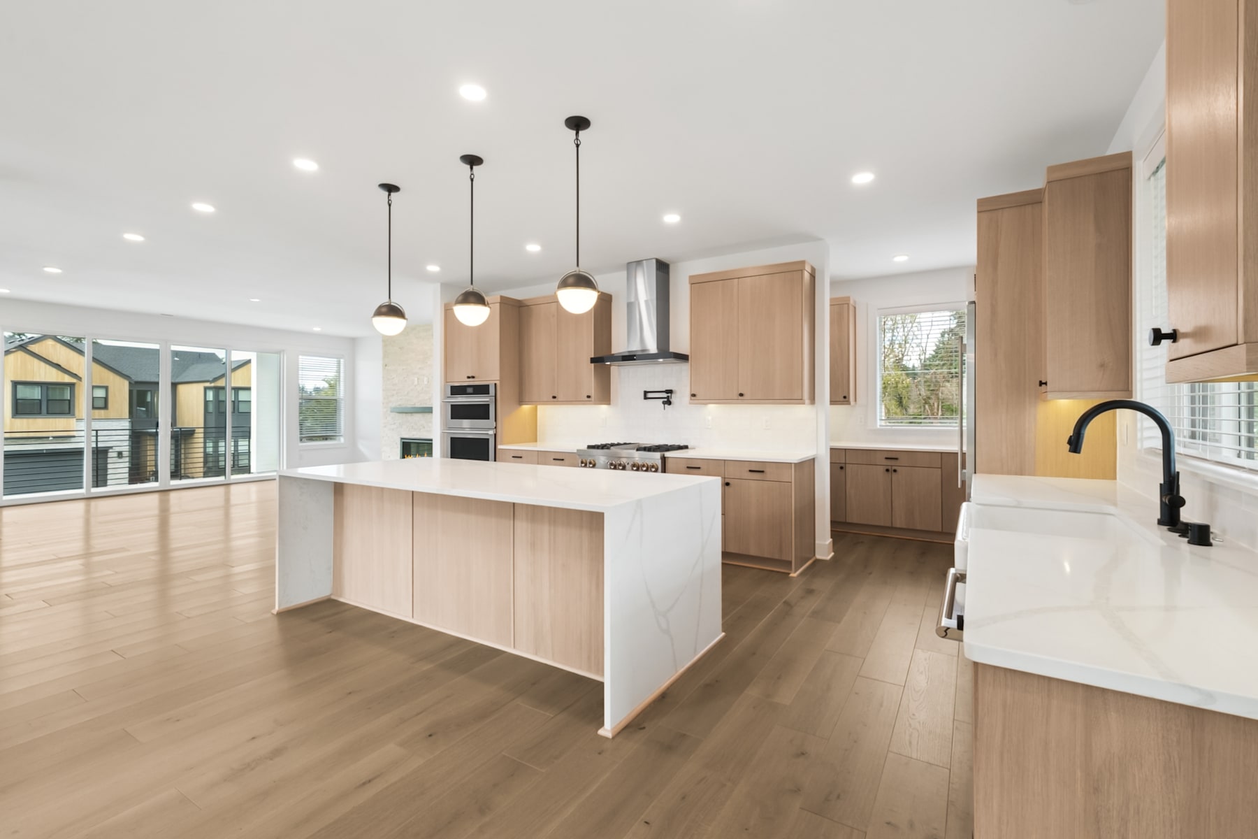 A modern, open-concept kitchen with light wood cabinetry, a large central island, and pendant lighting fixtures, set against a backdrop of hardwood floors and large windows.