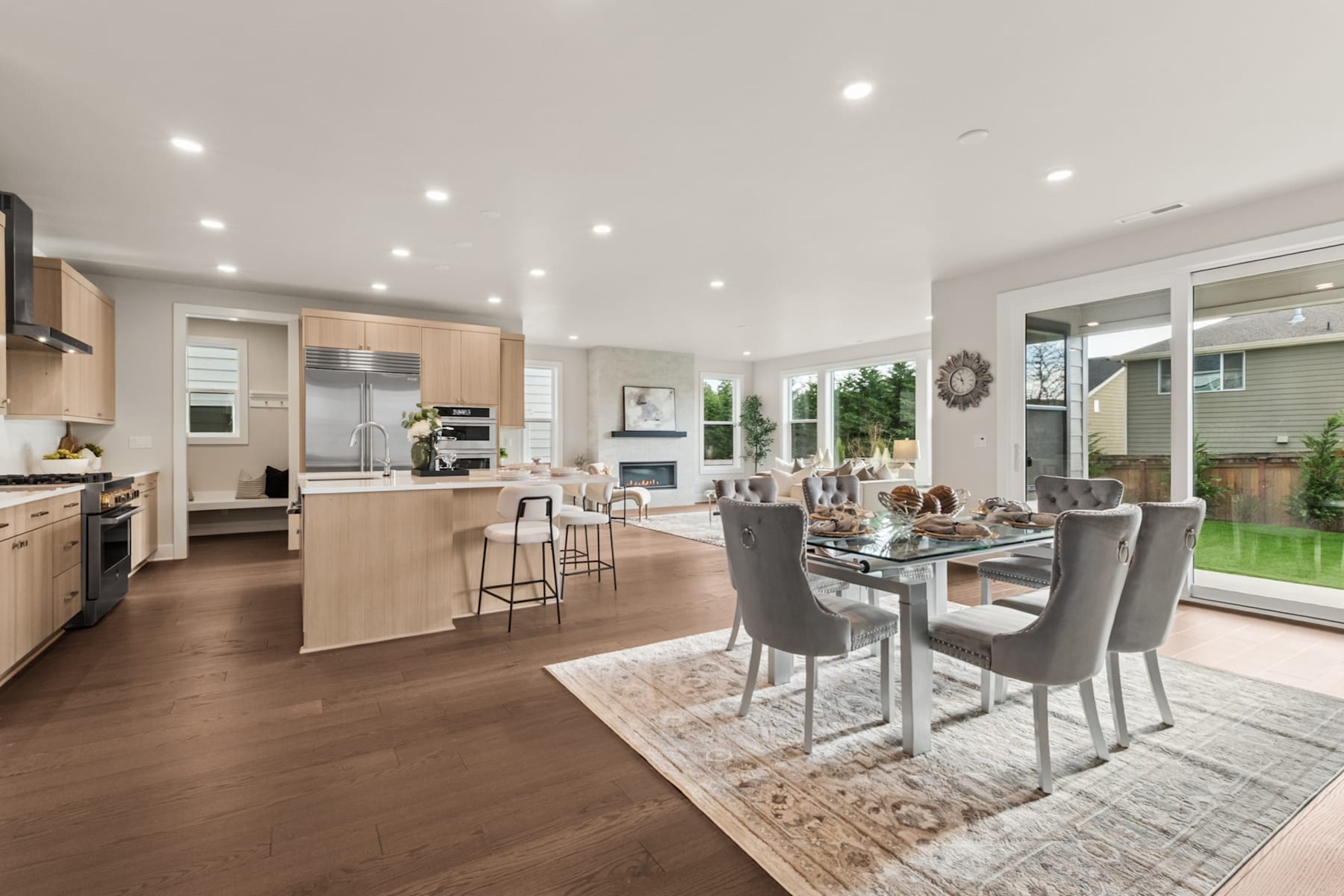 A modern and spacious kitchen and dining area with wooden floors, light-colored cabinets, and a large glass window overlooking a backyard.