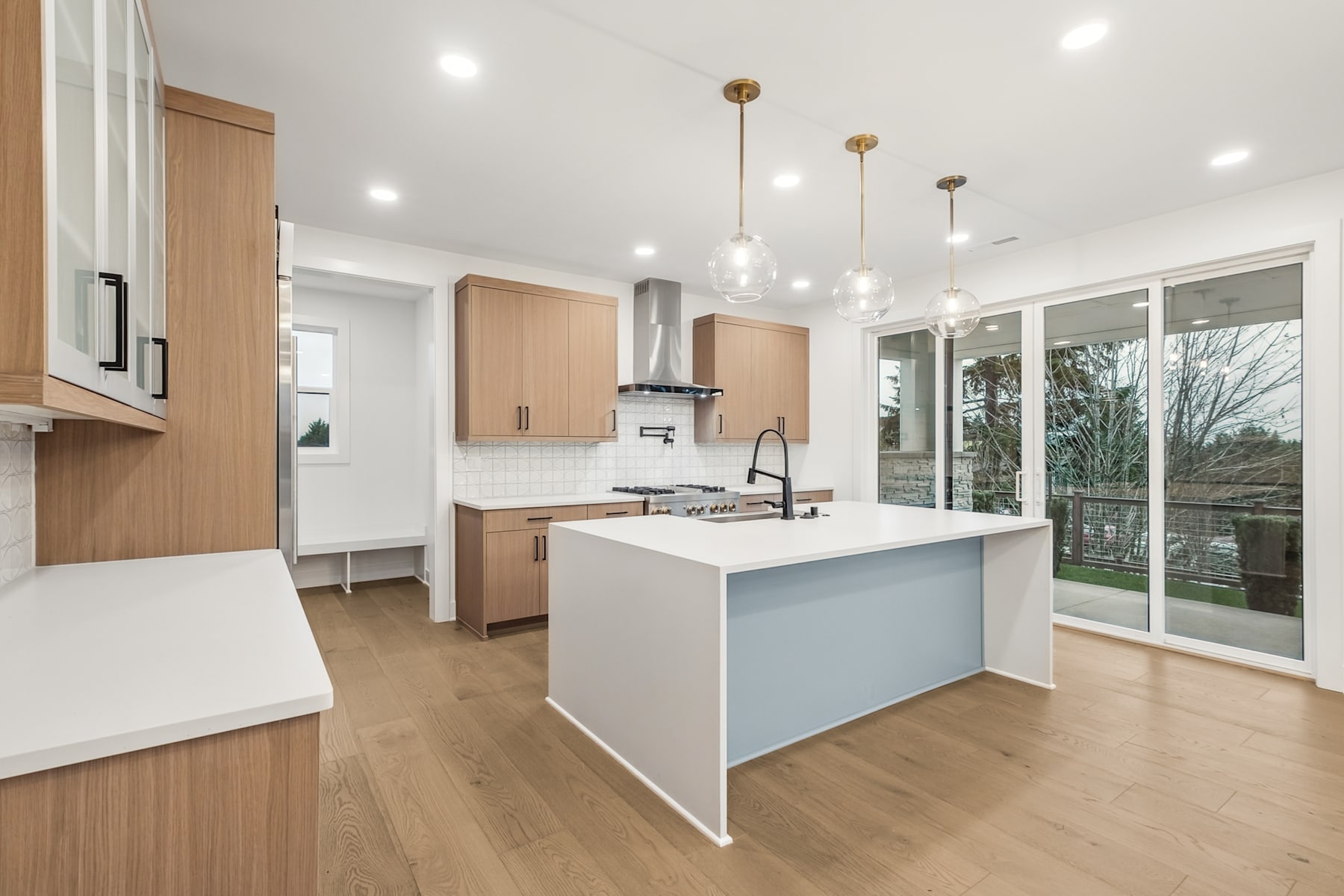 A modern, open-concept kitchen with light wood cabinets, a white island, and pendant lighting fixtures, set against a backdrop of large windows overlooking a wooded outdoor area.