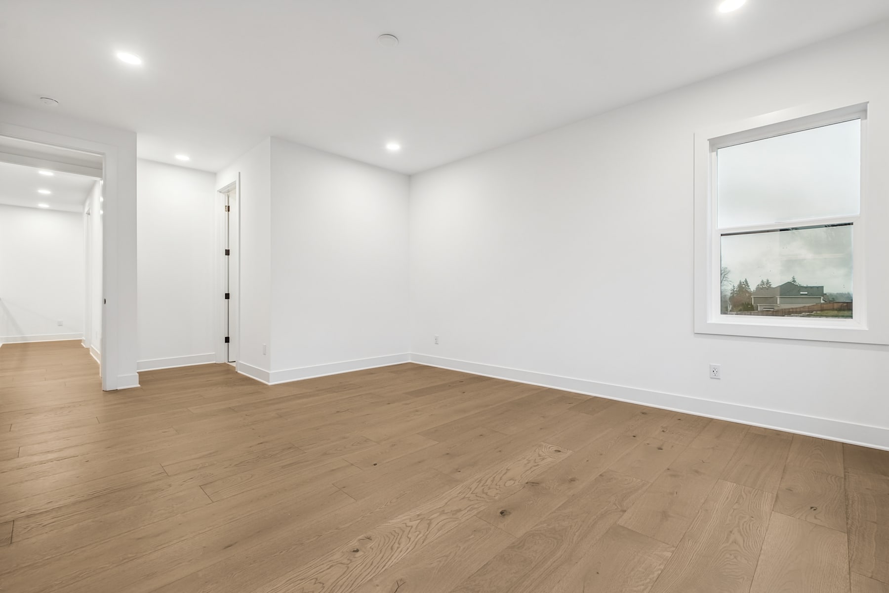A spacious, well-lit room with white walls and a large window displaying a scenic landscape, featuring hardwood floors and a doorway leading to another area.