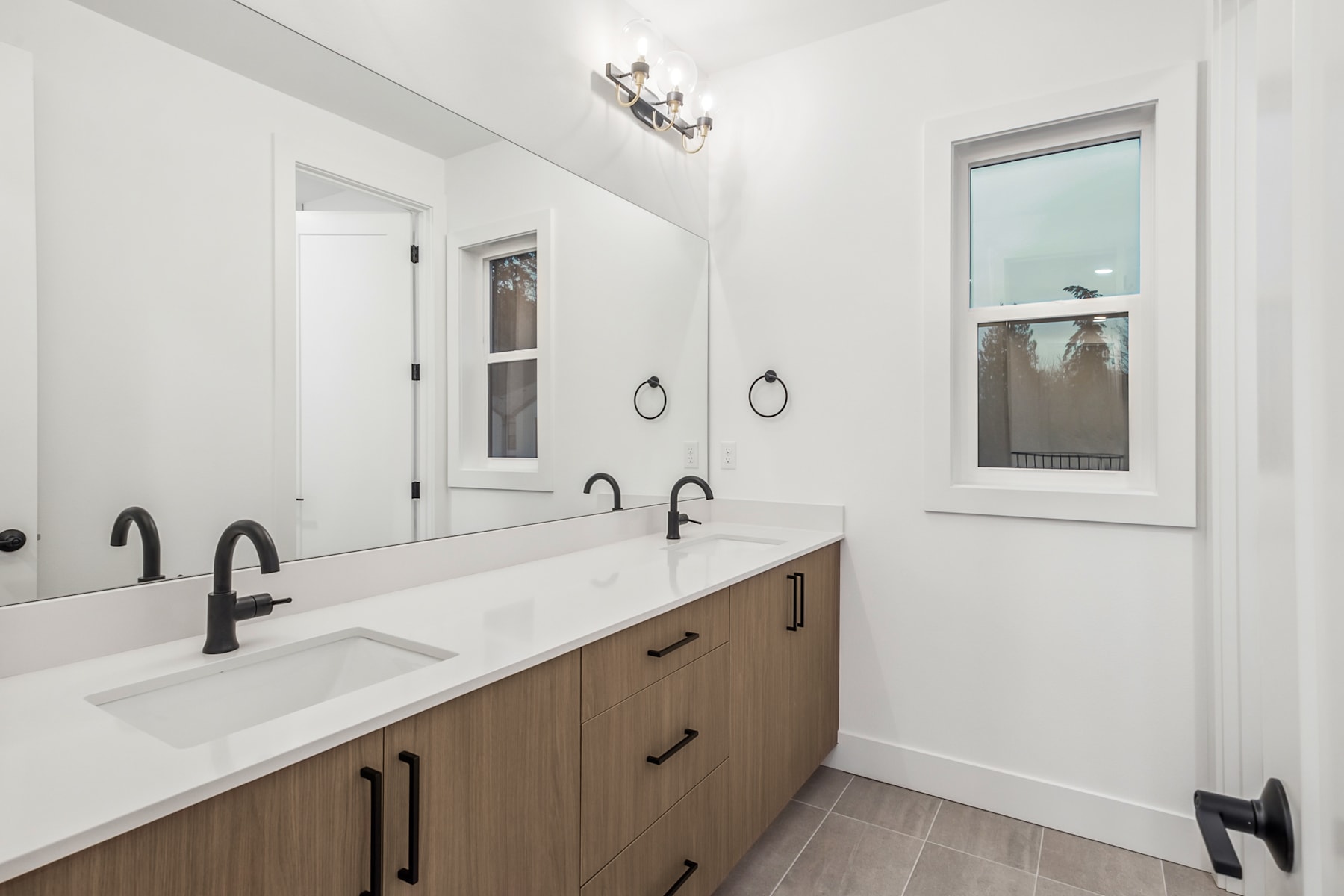 A modern and minimalist bathroom with a double vanity, white countertops, and dark hardware, set against a backdrop of white walls and a window.
