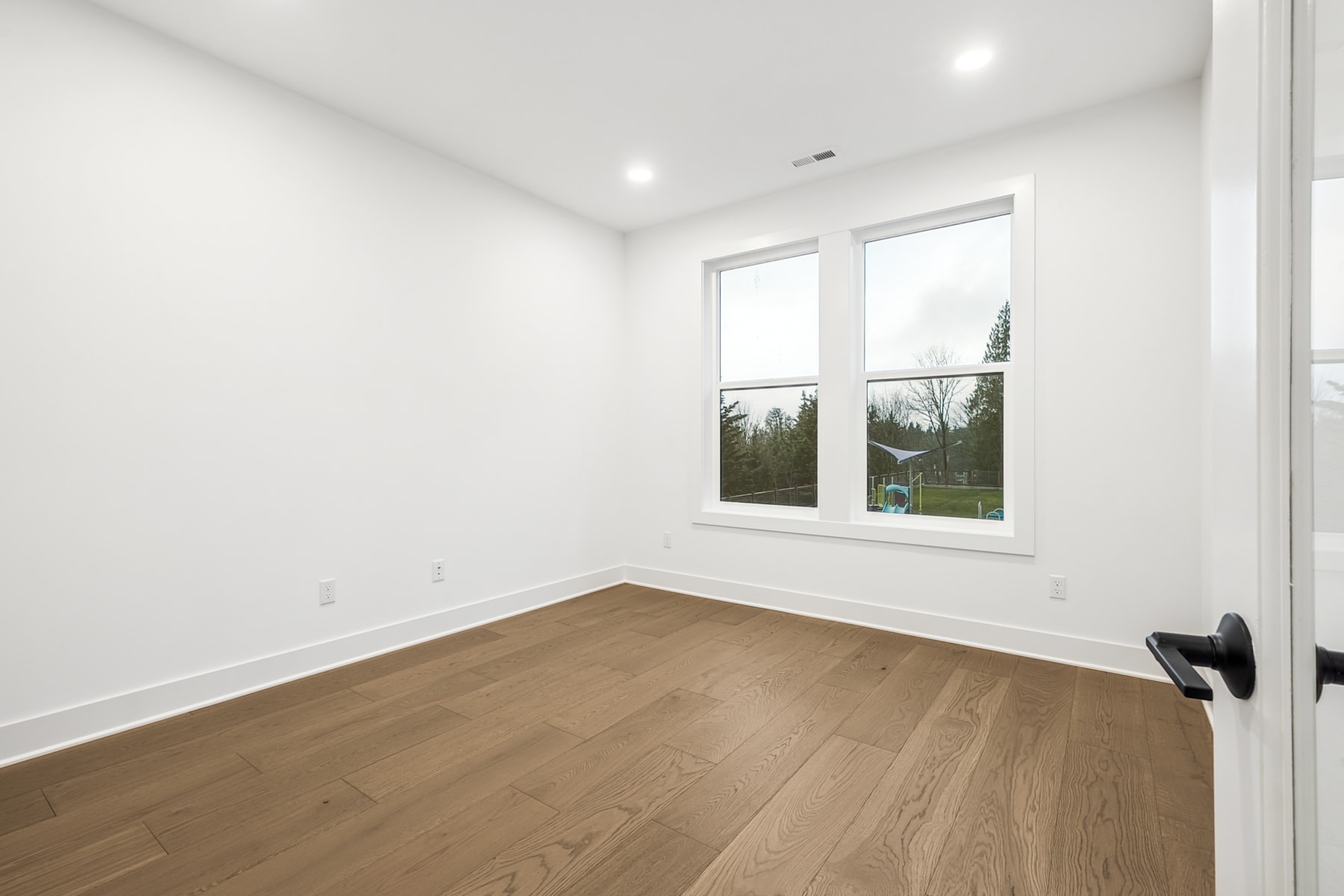 A bright, spacious room with large windows overlooking a wooded area, featuring a hardwood floor and a minimalist, clean design.