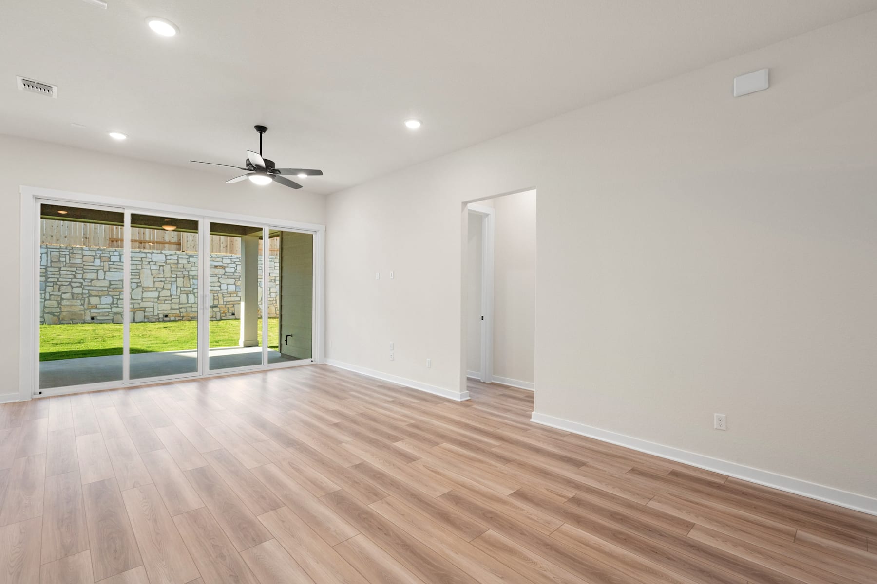 A spacious, well-lit room with hardwood floors, a ceiling fan, and a sliding glass door leading to a grassy outdoor area.
