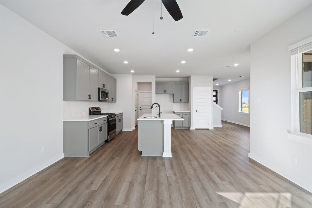 A modern, open-concept kitchen and living space with white walls, gray cabinets, and wood-look flooring, featuring a central island and various appliances.