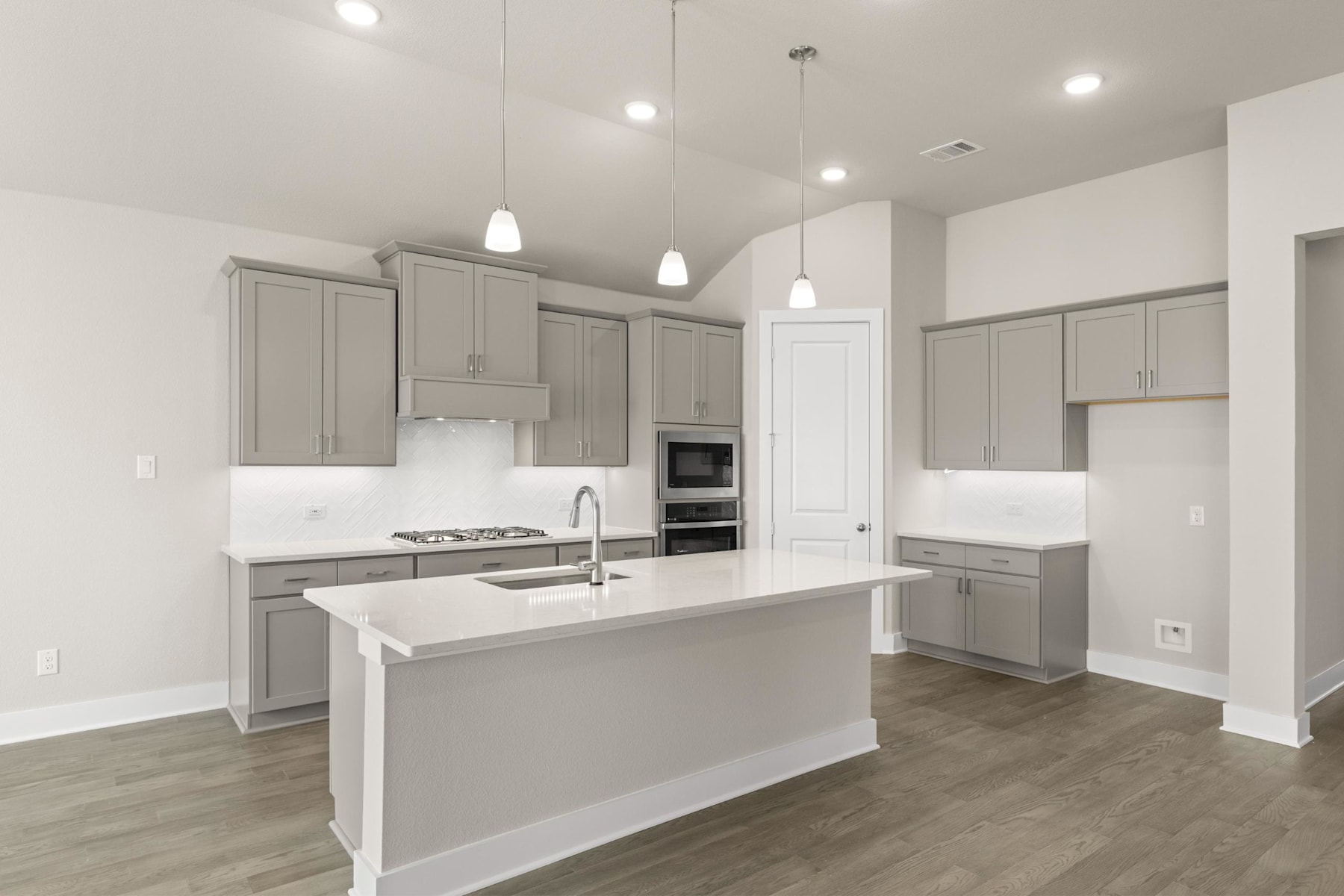 A modern, open-concept kitchen with gray cabinets, white countertops, and hardwood floors, featuring a central island and pendant lighting.