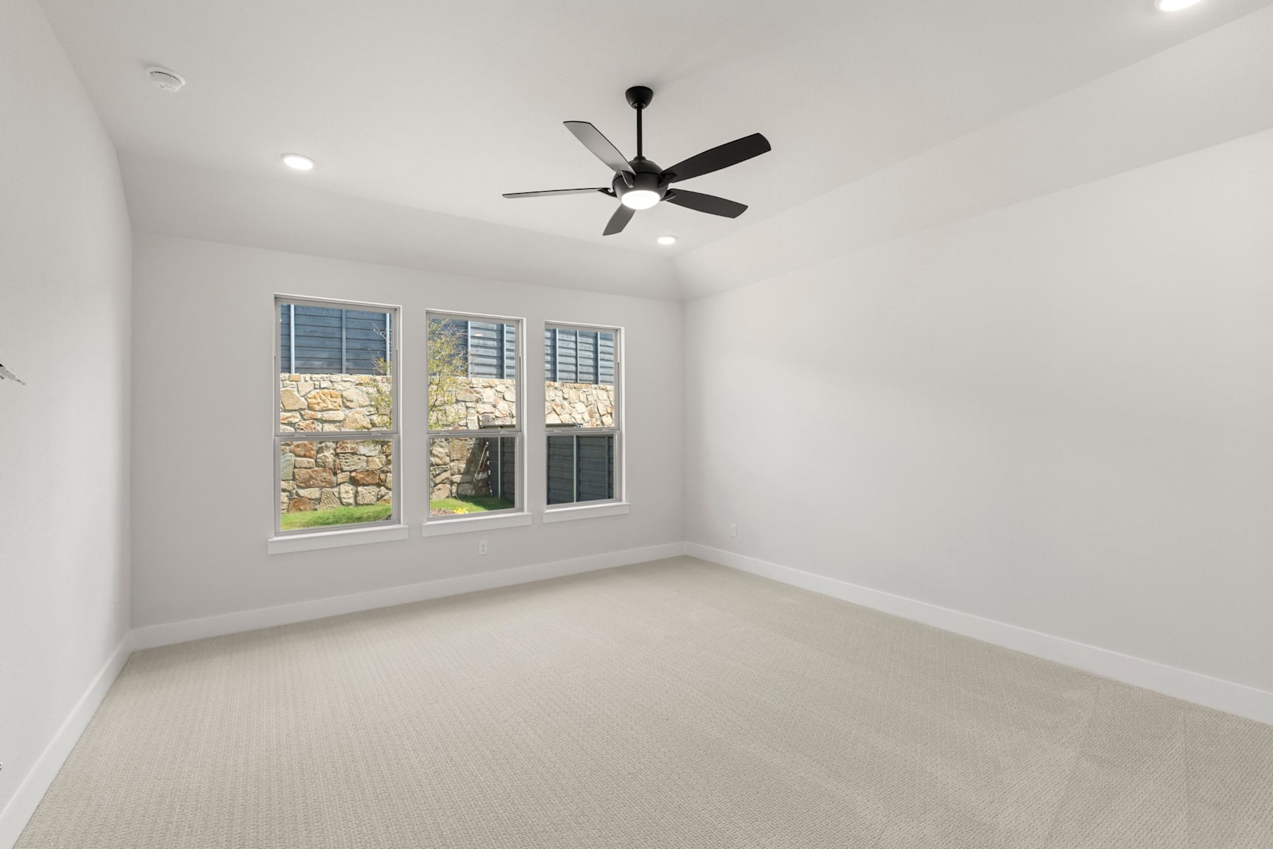 A bright, spacious room with a ceiling fan and large windows overlooking a stone exterior wall.