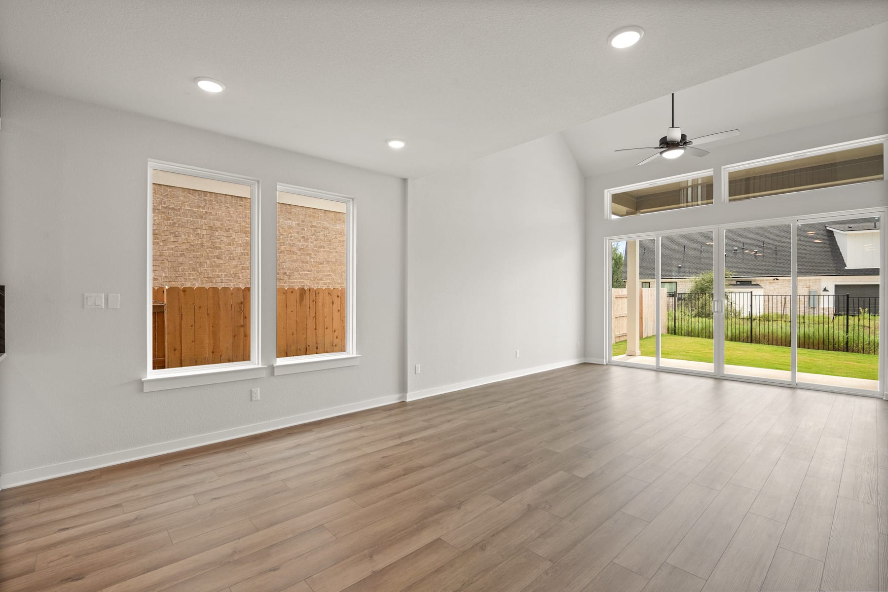 A spacious, well-lit room with wooden flooring, large windows, and a ceiling fan, overlooking a grassy outdoor area.