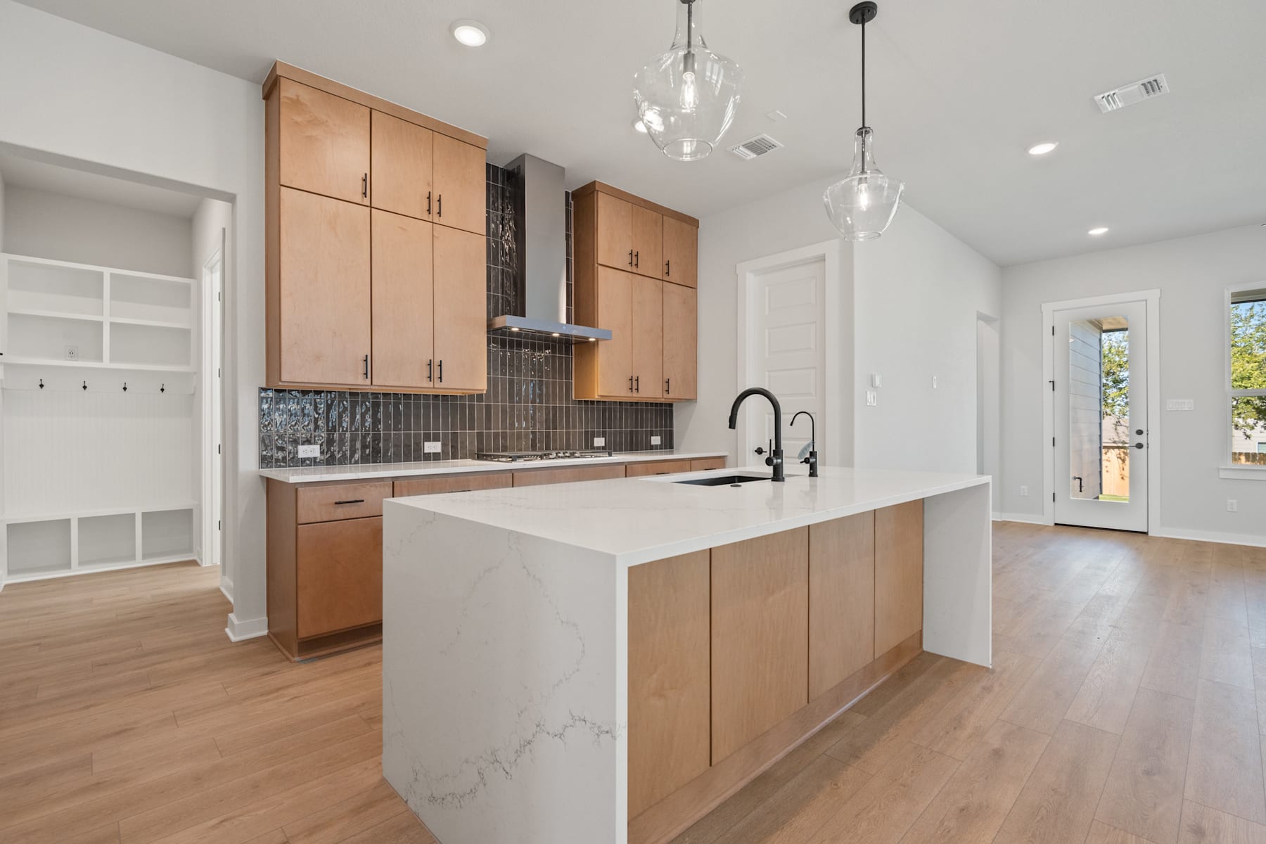A modern, open-concept kitchen with light wood cabinets, a white marble countertop, and pendant lighting fixtures, set against a backdrop of hardwood floors and a hallway leading to a bright, airy space.