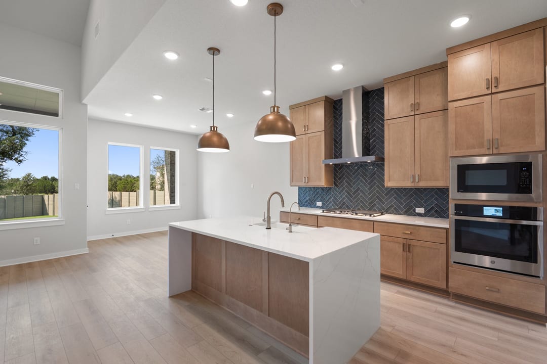 A modern and spacious kitchen with wooden cabinets, white countertops, and pendant lighting fixtures, set against a backdrop of large windows overlooking an outdoor area.