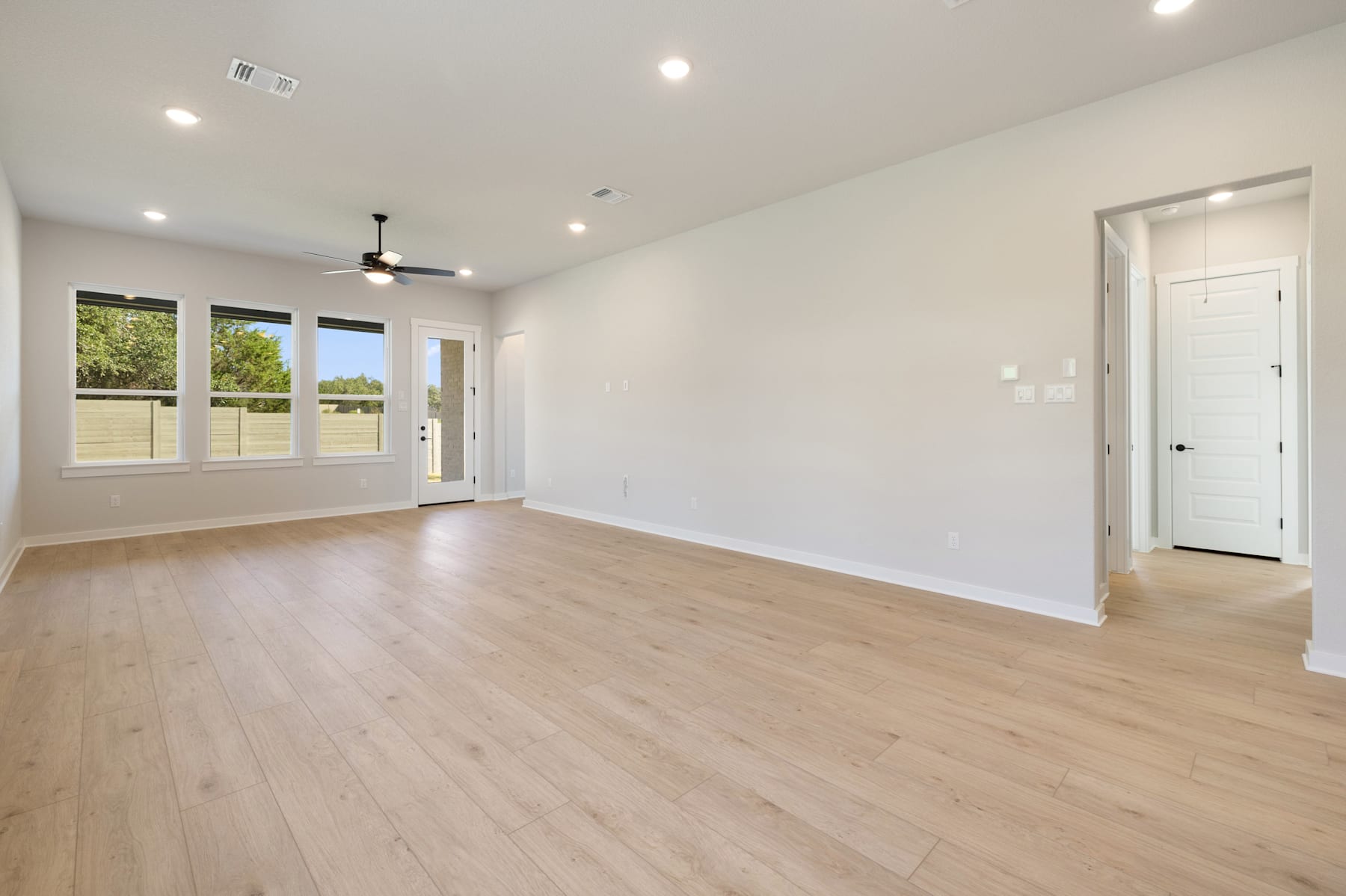A spacious, well-lit room with a hardwood floor, recessed lighting, and a ceiling fan. The room has large windows that provide a view of the outdoor scenery.