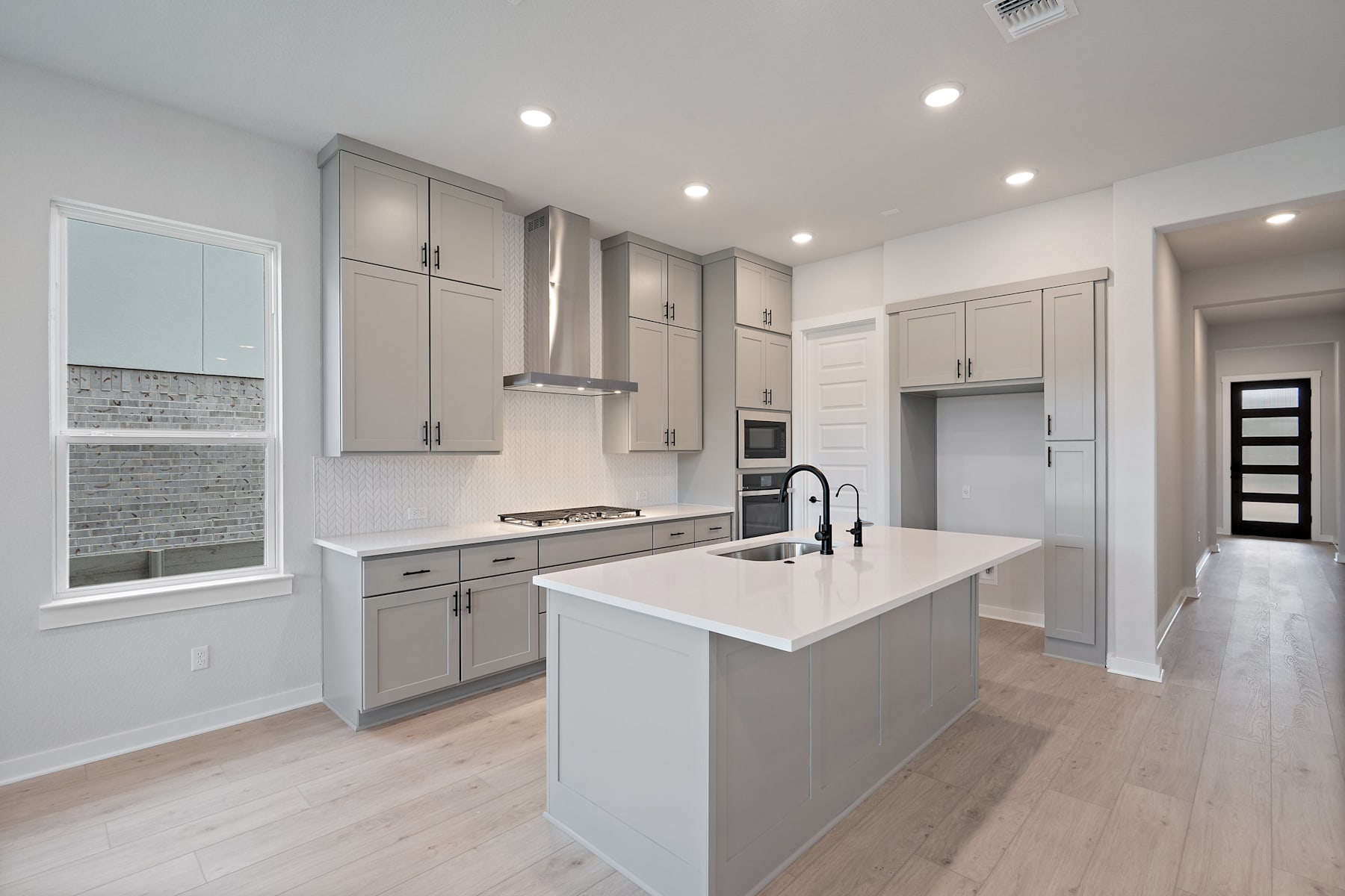 A modern, well-lit kitchen with white cabinets, gray countertops, and hardwood floors, featuring an island with a sink and a window overlooking the room.