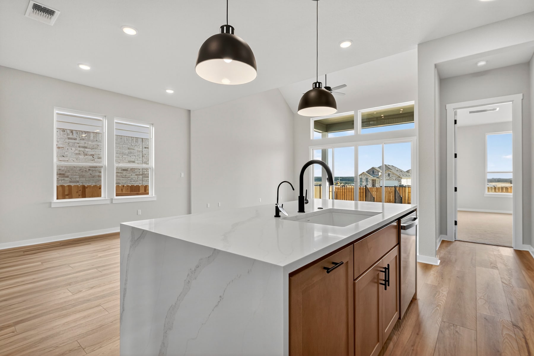 A modern and spacious kitchen with a large marble-topped island, wooden cabinets, and pendant lights hanging above, set against a backdrop of windows overlooking a residential neighborhood.