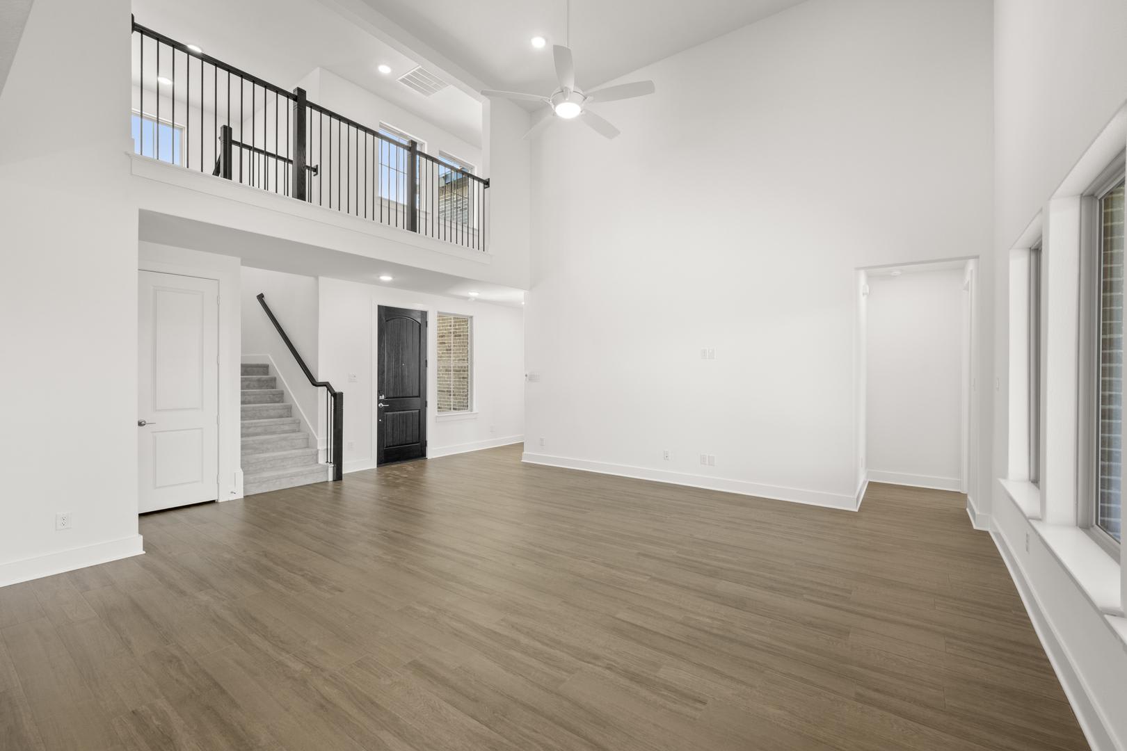 A spacious, open-concept room with white walls, a wooden floor, and a staircase leading to a second-floor balcony. The room appears to be a living or dining area, with a ceiling fan and large windows providing natural light.