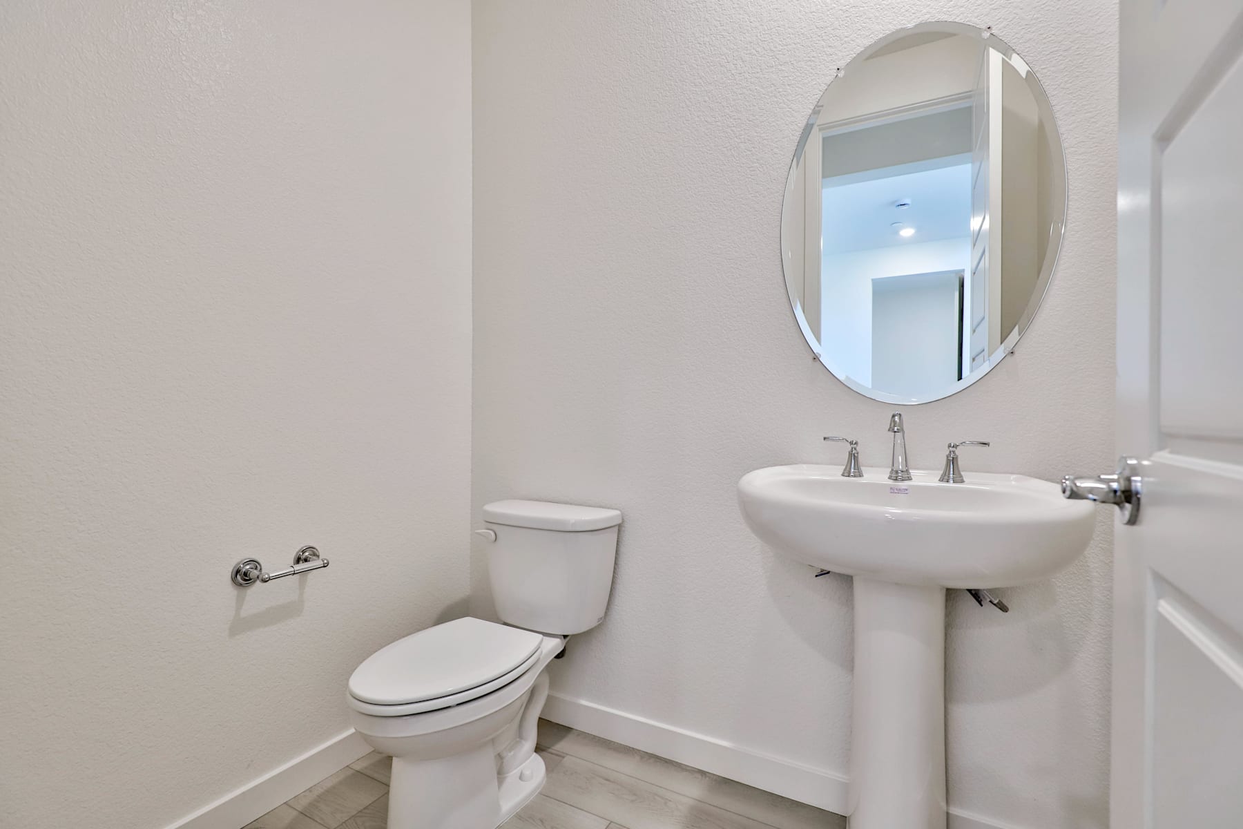 A clean, well-lit bathroom with a white toilet, a pedestal sink, and an oval mirror on the wall.