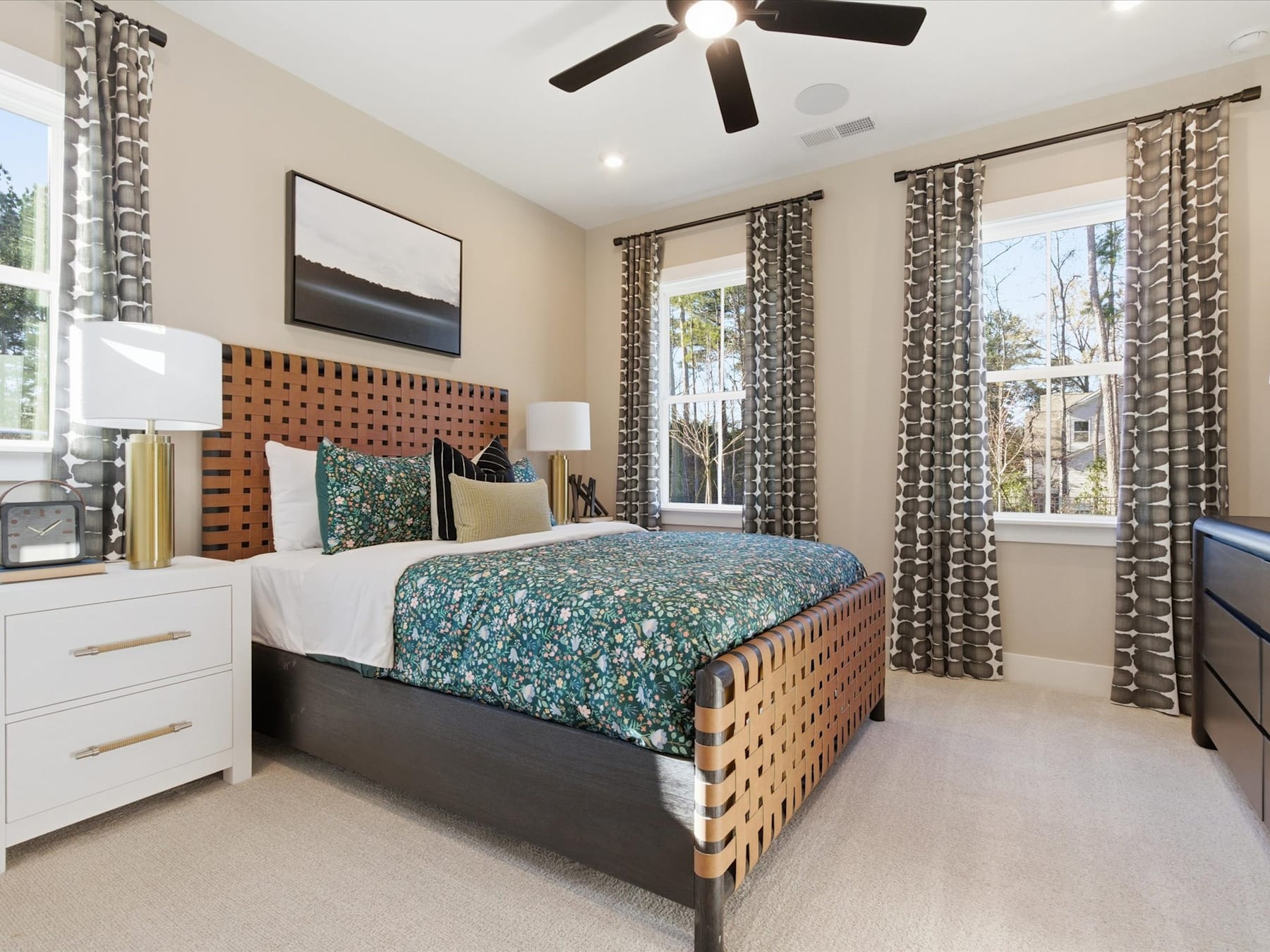 A cozy and well-furnished bedroom with a patterned bedspread, wooden bed frame, and curtains, along with a ceiling fan and a window providing natural light.