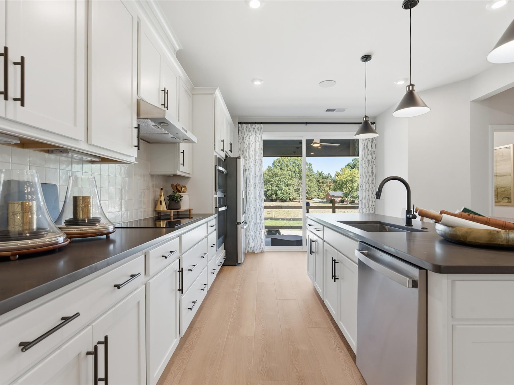 A modern, well-lit kitchen with white cabinets, dark countertops, and stainless steel appliances, overlooking a scenic outdoor view through a glass door.