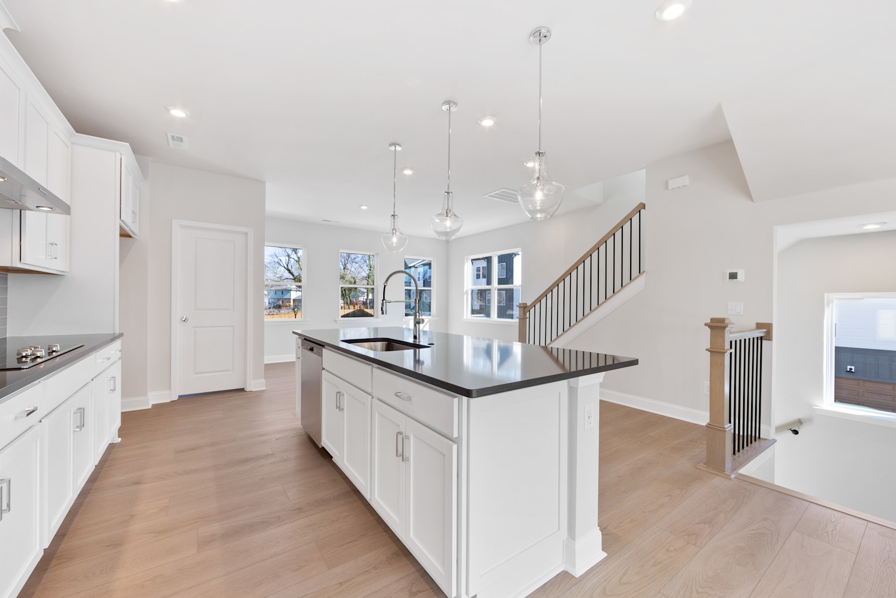 A spacious and modern kitchen with white cabinets, a large island, and hardwood floors, with a staircase and open concept layout visible in the background.