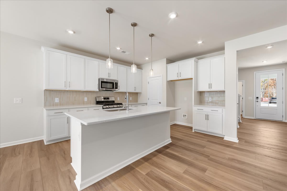 A modern, open-concept kitchen with white cabinets, stainless steel appliances, and a central island with a light-colored countertop, set against a hardwood floor.