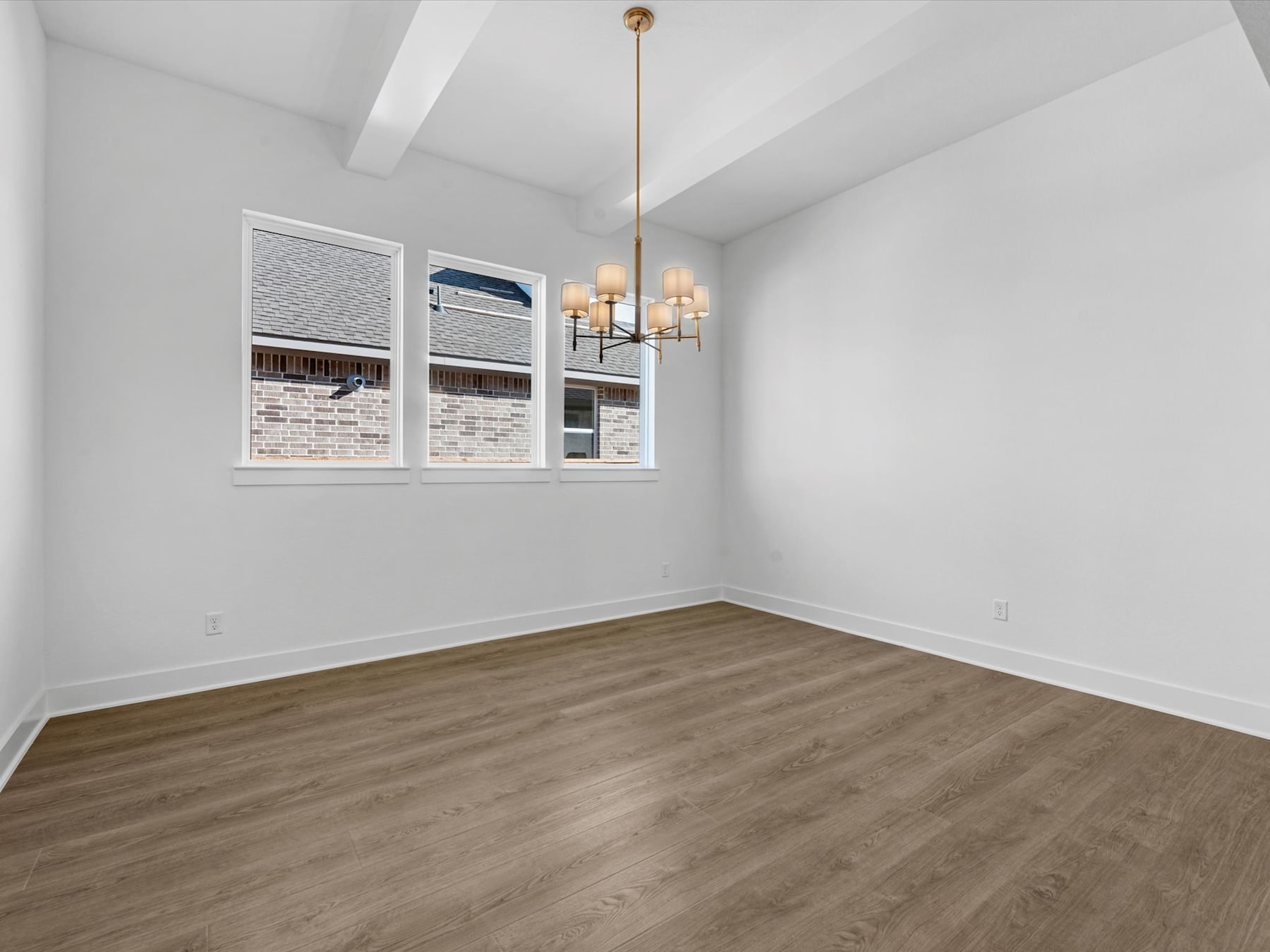 A bright, minimalist room with white walls, a wooden floor, and a chandelier hanging from the ceiling. The room appears to be empty, with built-in shelves on one wall.