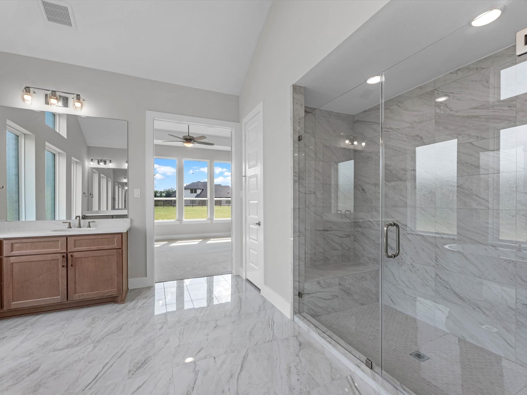 A spacious and modern bathroom with a large glass shower enclosure, a vanity with wooden cabinets, and a view of the outdoor landscape through the windows.