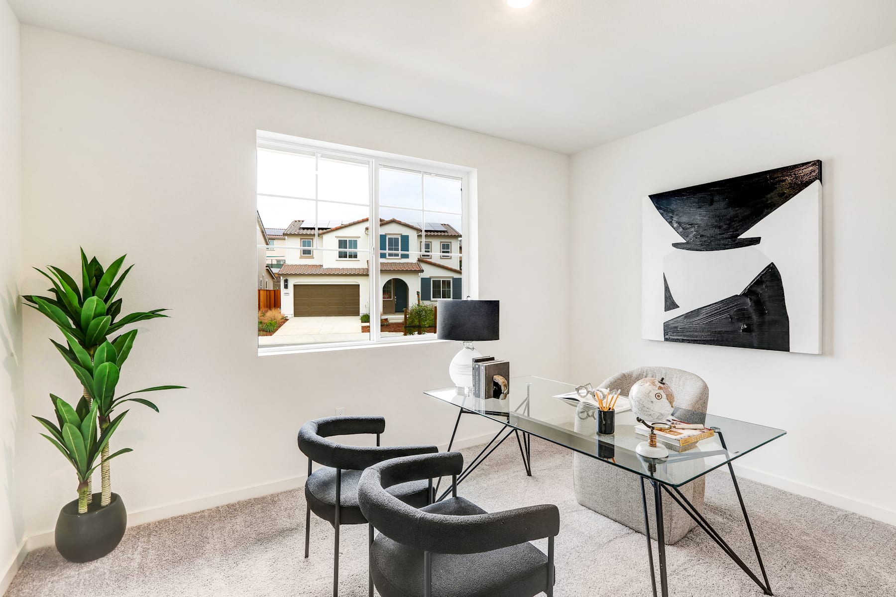 A modern and minimalist home office space with a glass desk, black chairs, and abstract artwork on the walls, set against the backdrop of a residential neighborhood visible through the window.