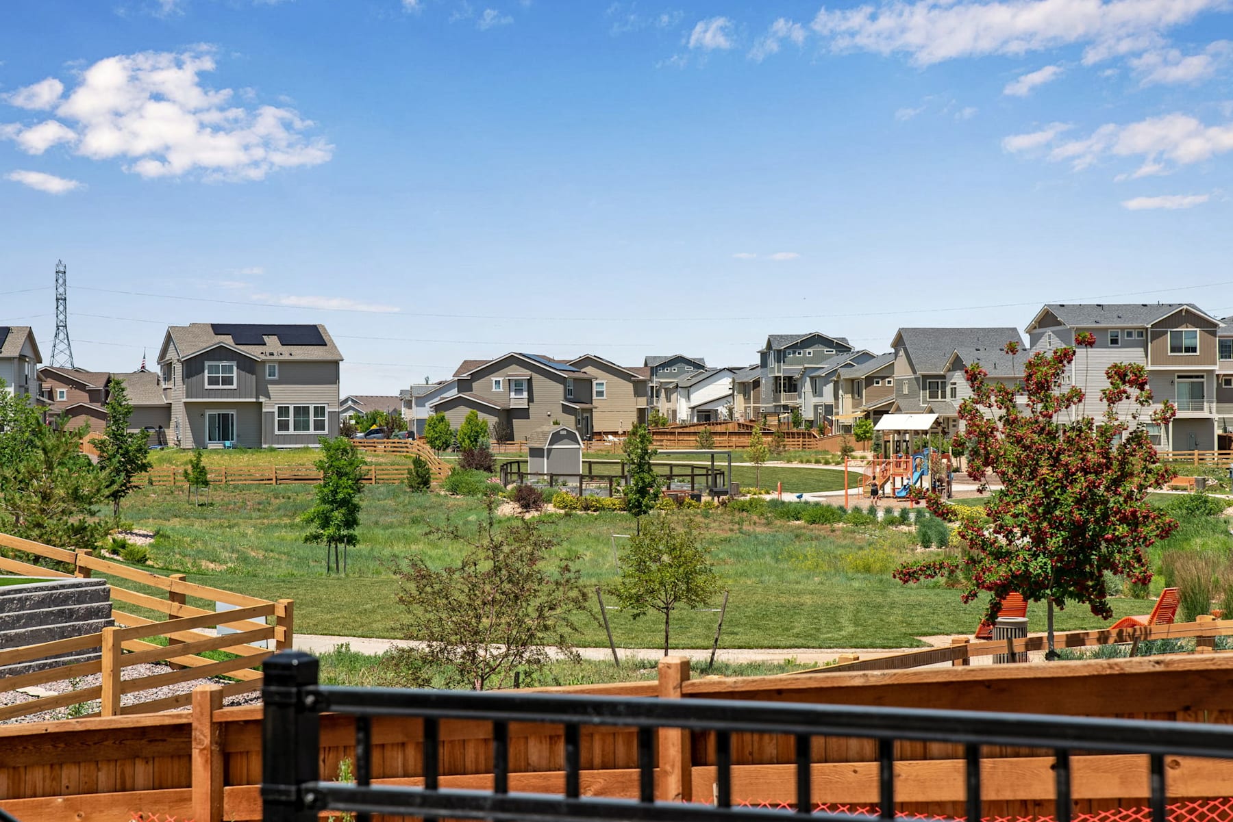A residential neighborhood with various houses, some with solar panels, surrounded by a grassy area with trees and a wooden fence in the foreground.