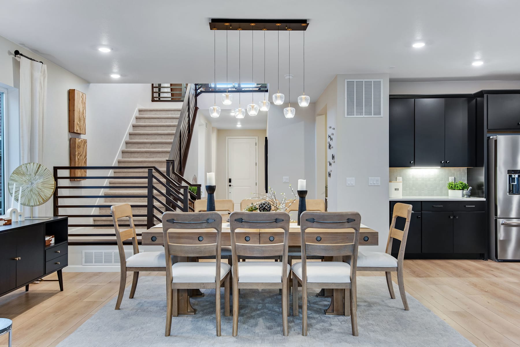 A modern and open-concept dining area with a wooden table and chairs, pendant lighting, and a staircase leading to an upper level in the background.