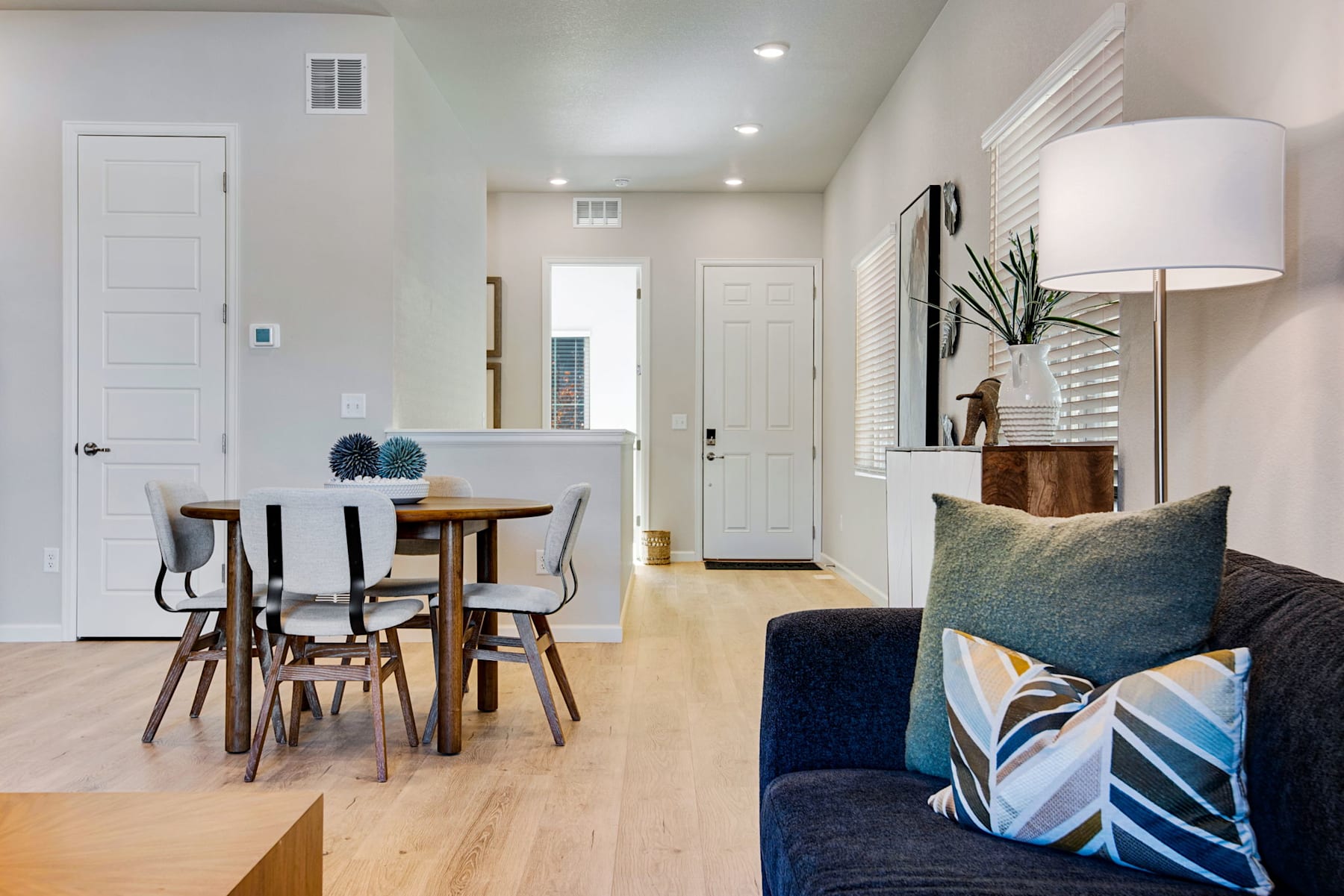 A modern and minimalist dining area with a wooden table and chairs, adjacent to a cozy living space with a patterned throw pillow on a dark sofa, and a hallway leading to other rooms in the home.