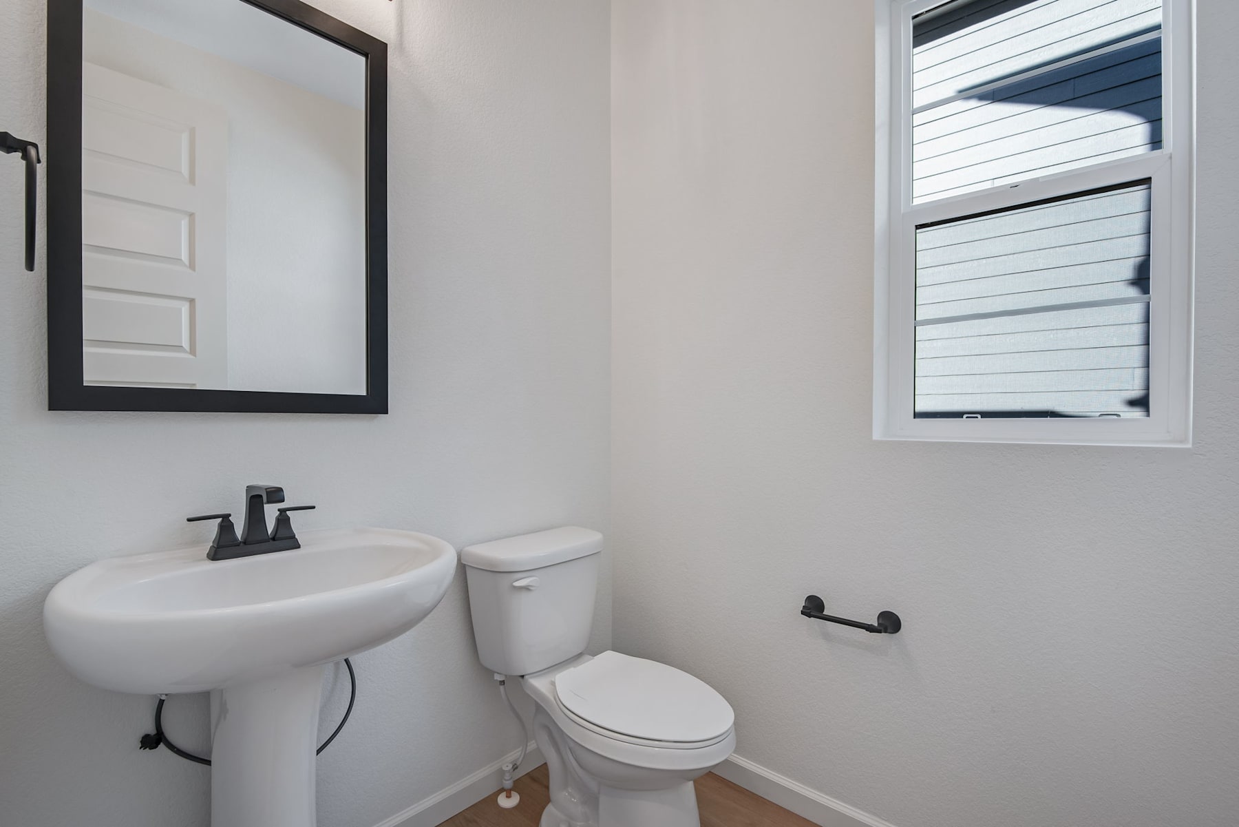 A modern, minimalist bathroom with a white pedestal sink, a toilet, and a framed mirror on the wall. The room has a clean, simple design with neutral colors and natural lighting from the window.