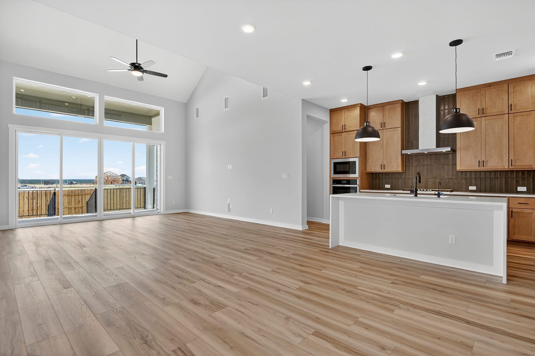 A modern, open-concept kitchen and living room with wooden cabinets, hardwood floors, and large windows providing natural light.