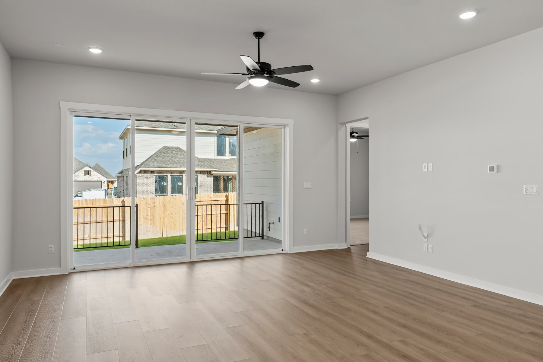 A spacious and bright room with a sliding glass door leading to a backyard, featuring a ceiling fan and hardwood flooring.