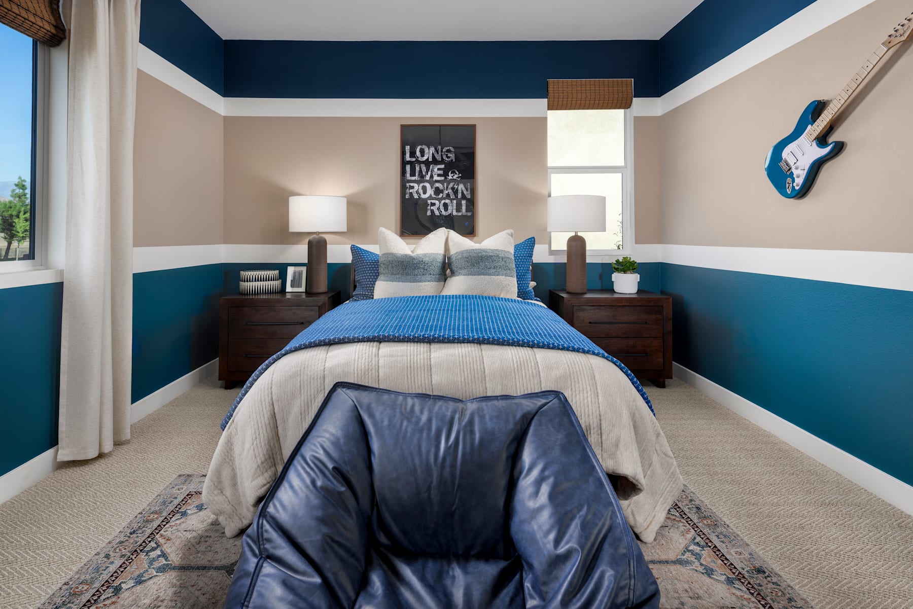A cozy and stylish bedroom with a blue and beige color scheme, featuring a bed with a blue comforter, wooden nightstands, and a decorative guitar on the wall.