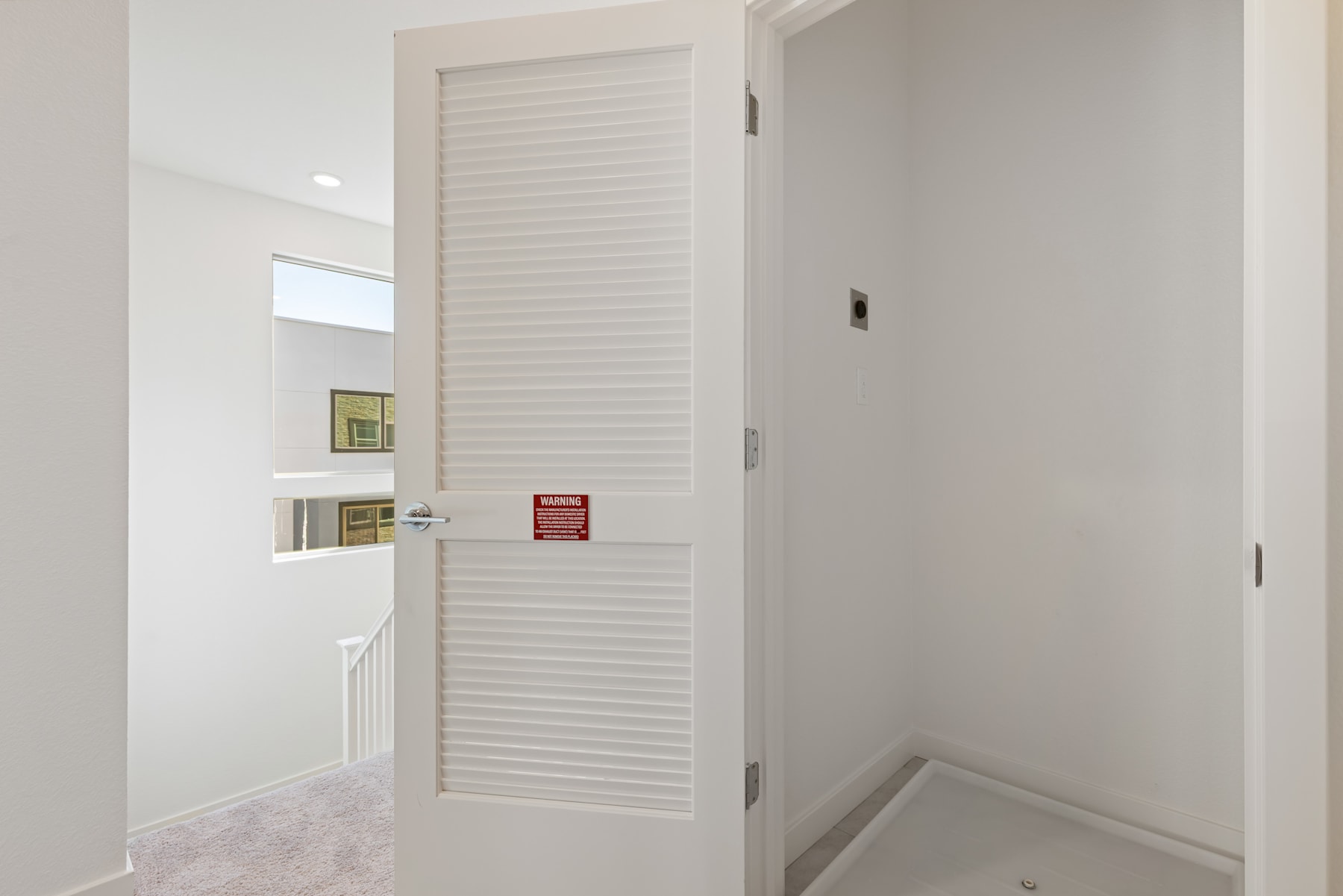 A white louvered door stands in the foreground, leading to a hallway with white walls and a small shelf on the side.