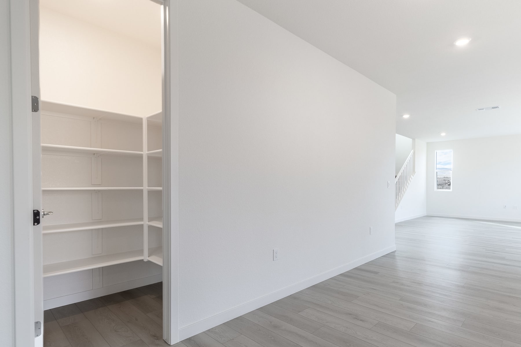 A white, minimalist hallway with an open closet or storage space on the left, and a hardwood floor leading to a brightly lit room in the background.