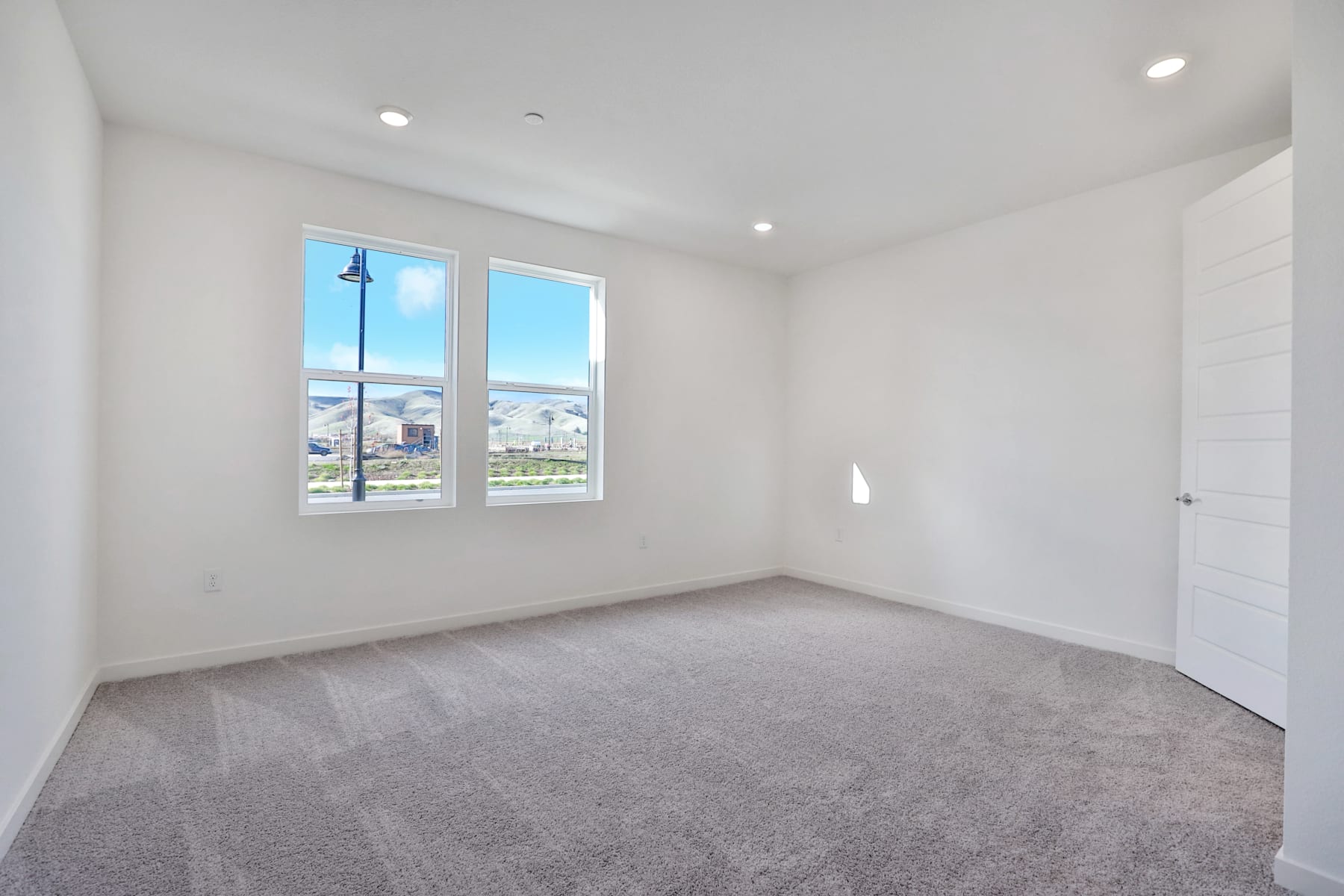 A spacious, well-lit room with large windows overlooking a scenic outdoor landscape, featuring a clean and minimalist interior design with gray carpeting and white walls.