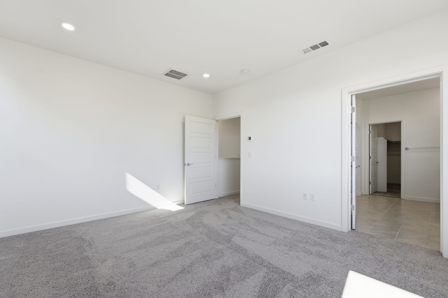 An empty, minimalist room with white walls, a gray tiled floor, and a doorway leading to another room.