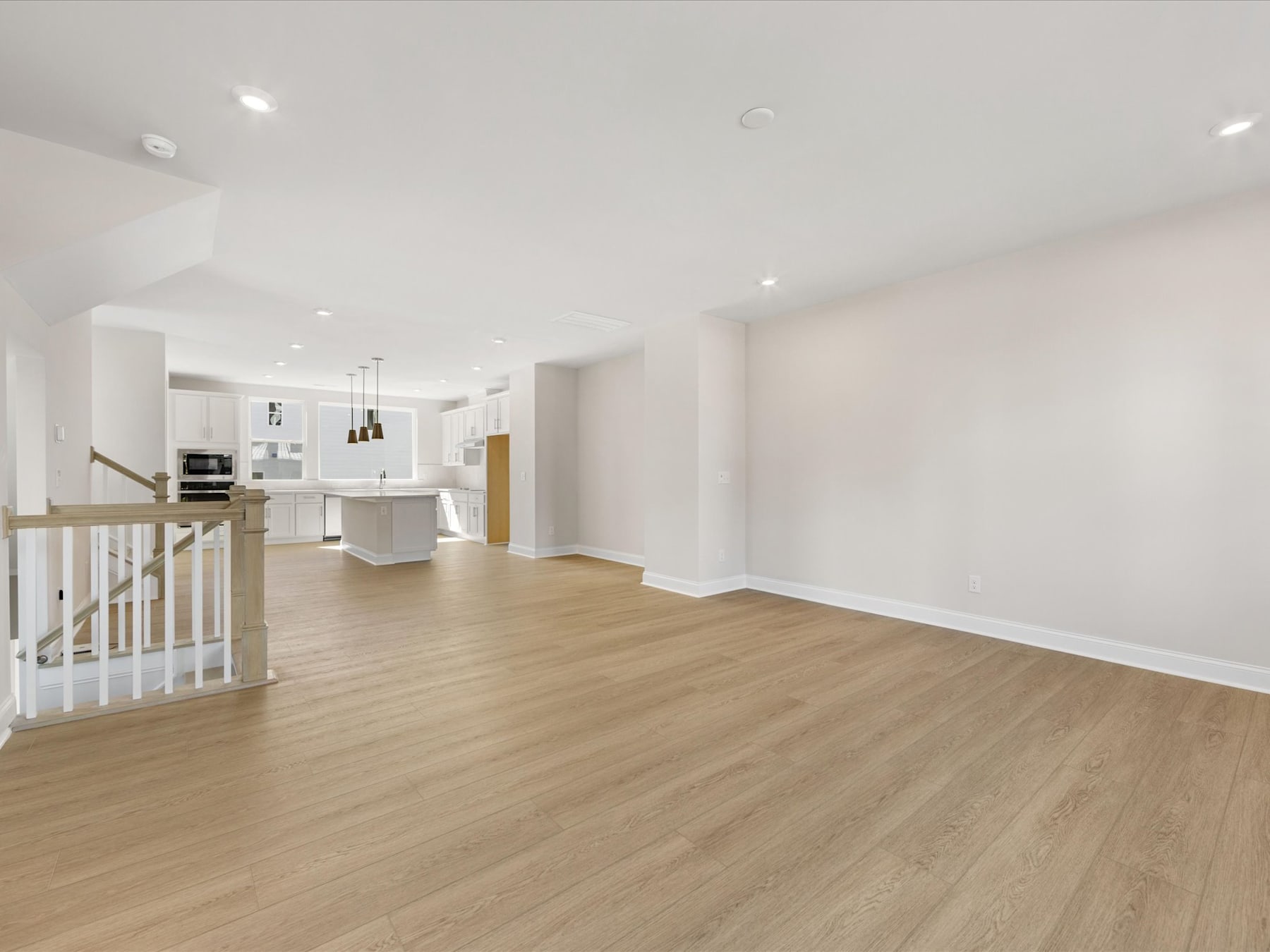 A spacious, open-concept living area with a white wall, hardwood floors, and a kitchen visible in the background.