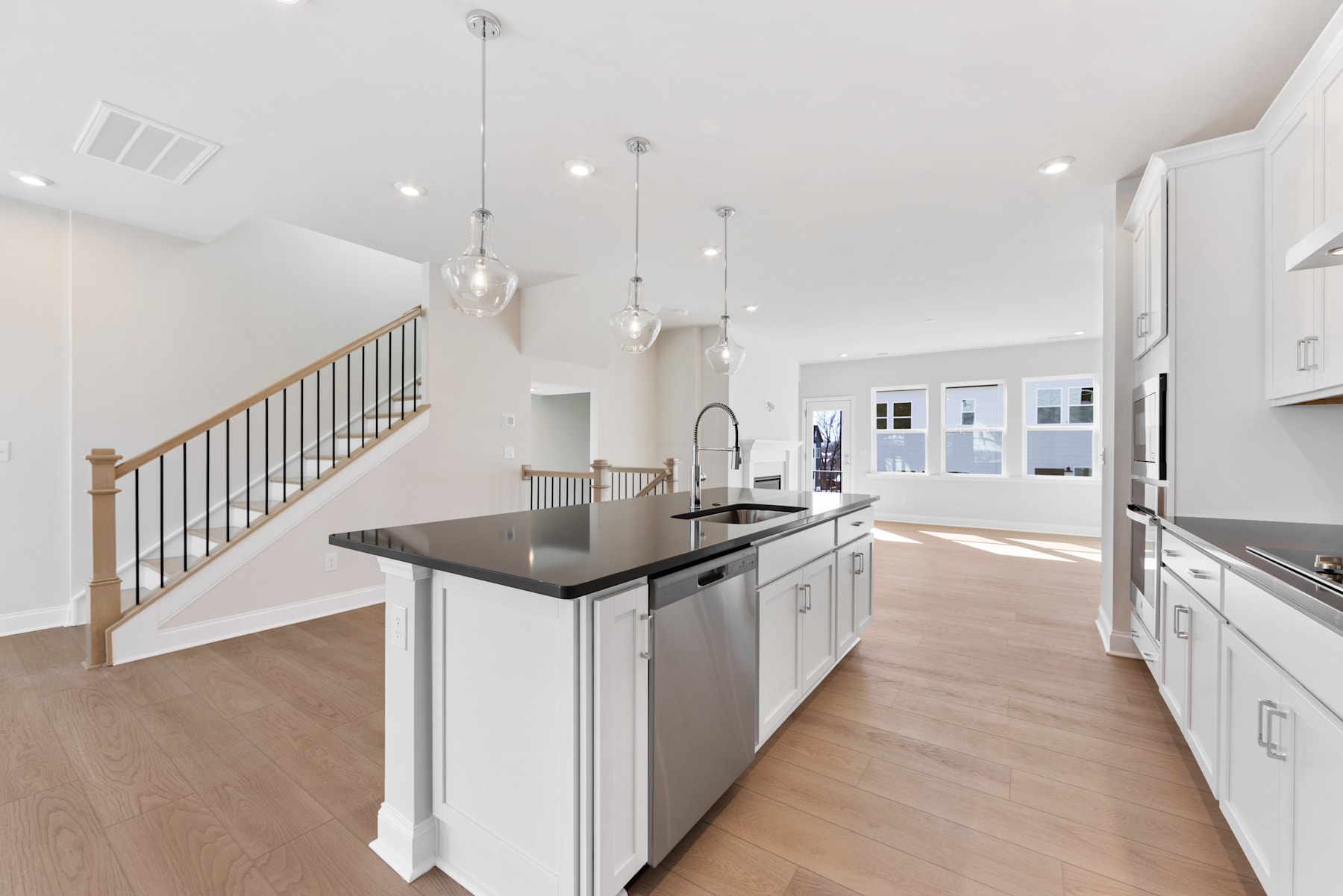 A modern, open-concept kitchen with white cabinets, stainless steel appliances, and pendant lighting, leading into a bright and airy living space with a wooden staircase.
