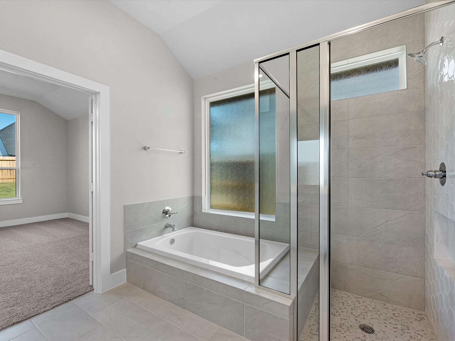 A spacious and modern bathroom with a large bathtub, tiled walls, and a glass-enclosed shower area, set against a backdrop of a carpeted bedroom.