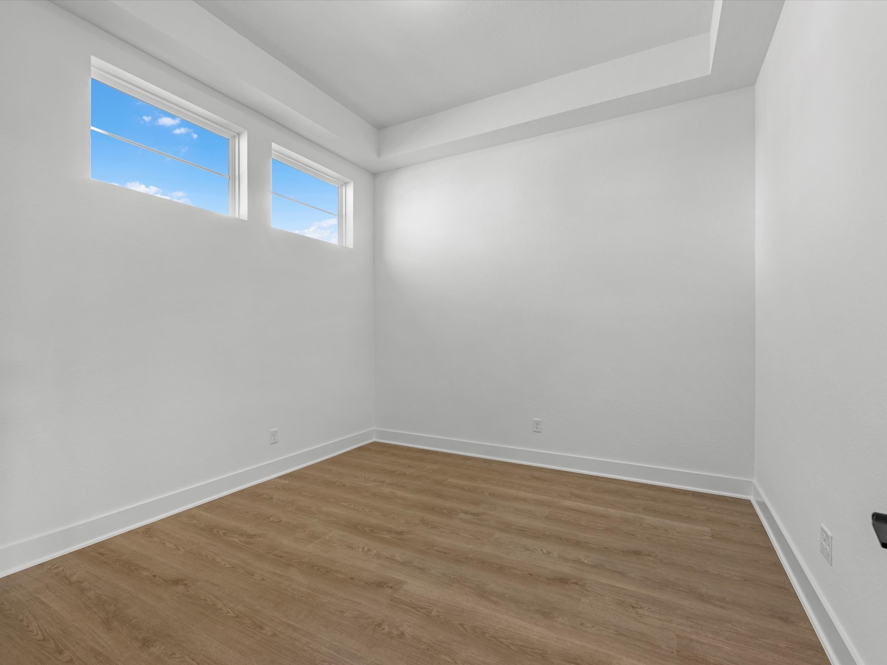 A bright, empty room with white walls and a wooden floor, with two windows providing natural light and a view of the sky.