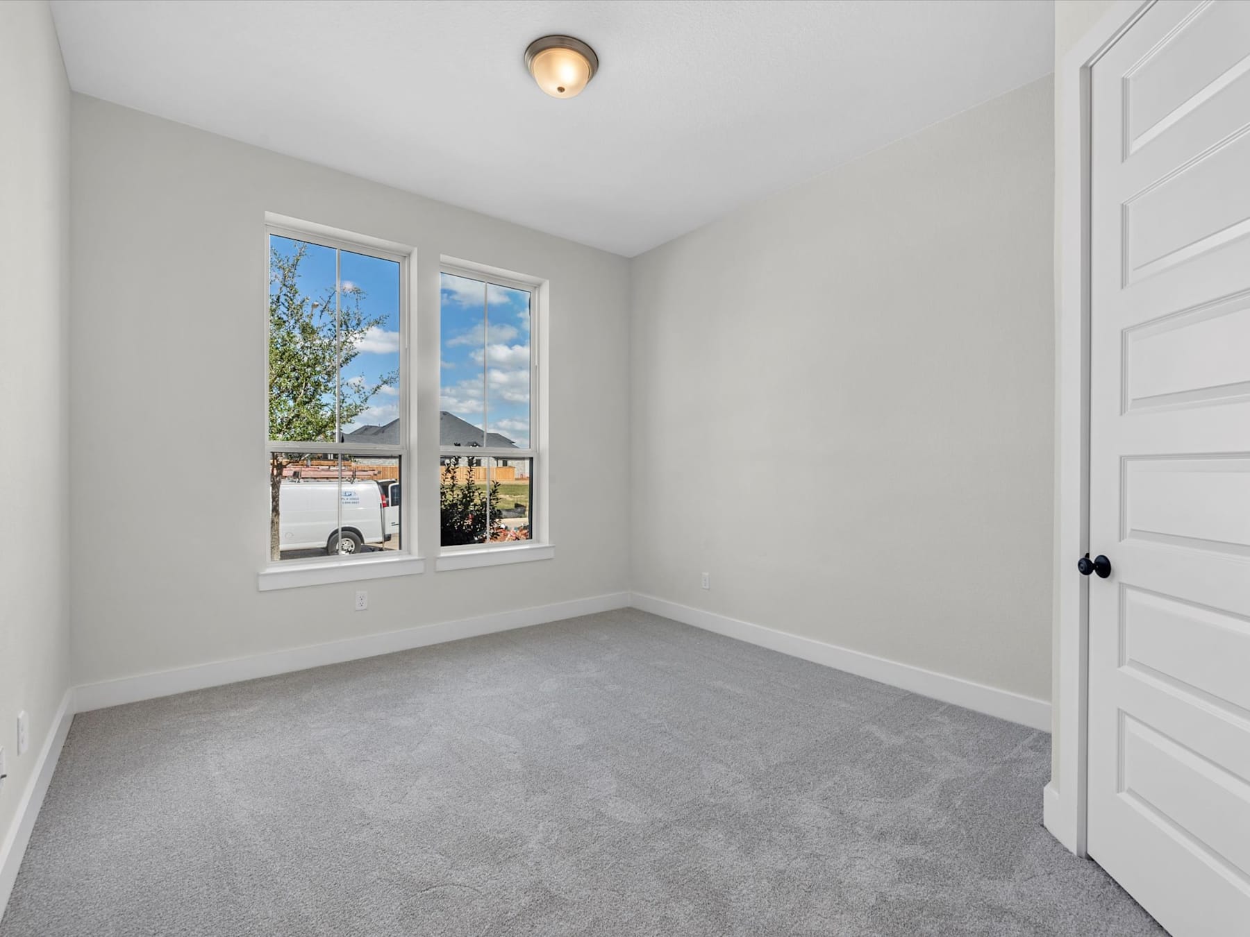 A bright and spacious room with large windows overlooking the outdoors, featuring a neutral-toned interior and a closed door leading to another area.