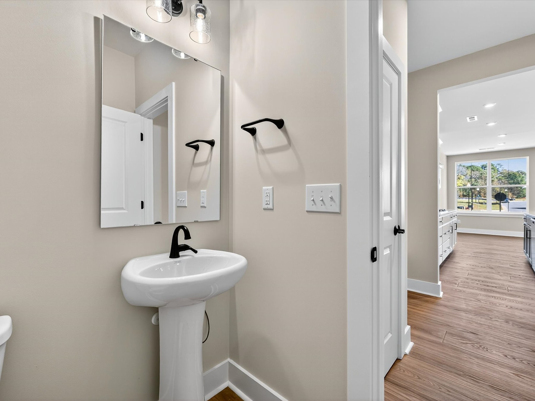 A modern and minimalist bathroom with a white pedestal sink, black fixtures, and a mirror on the wall. The room has a neutral color scheme and appears to be part of a larger living space with a visible hallway in the background.