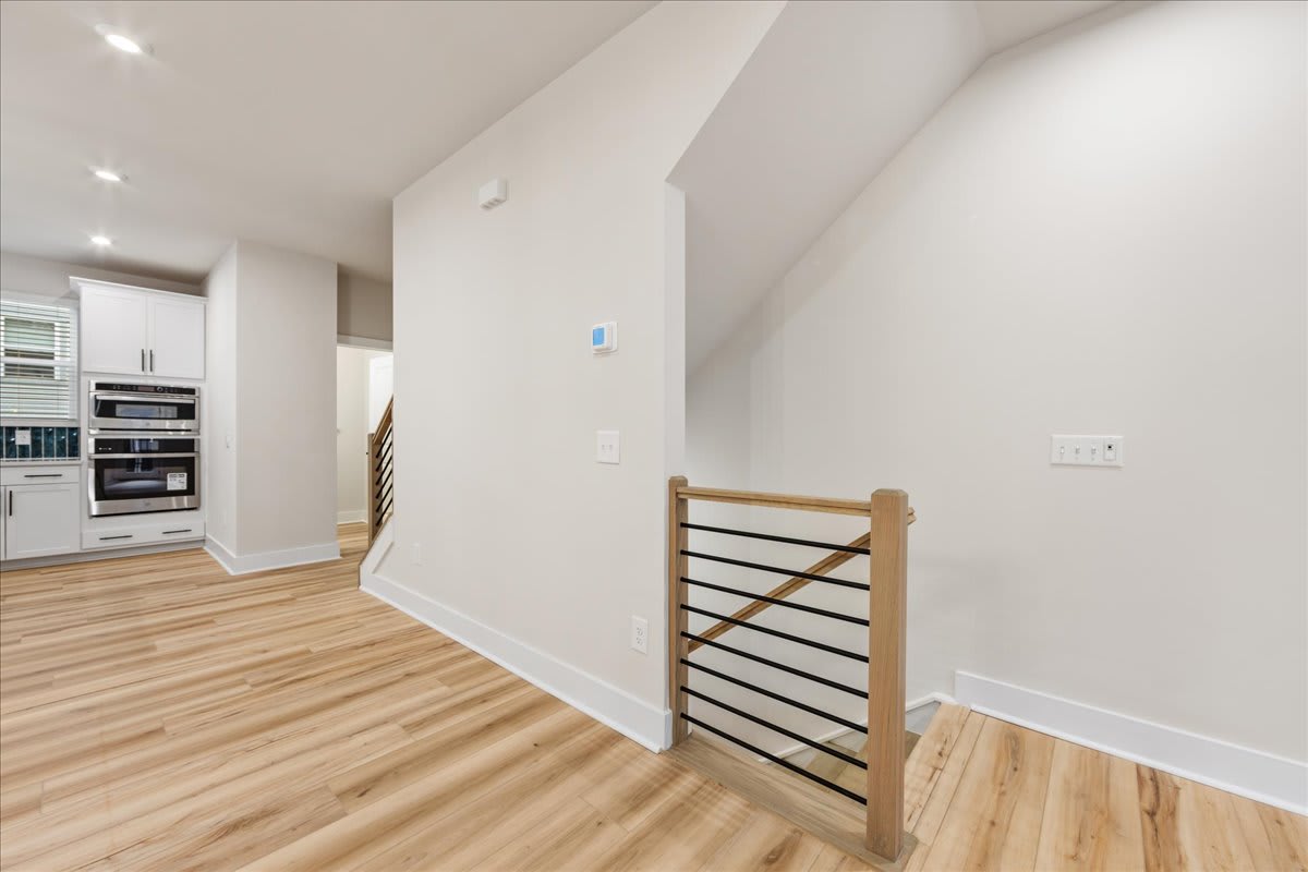 A bright and airy interior space with a wooden staircase railing in the foreground and a kitchen visible in the background.