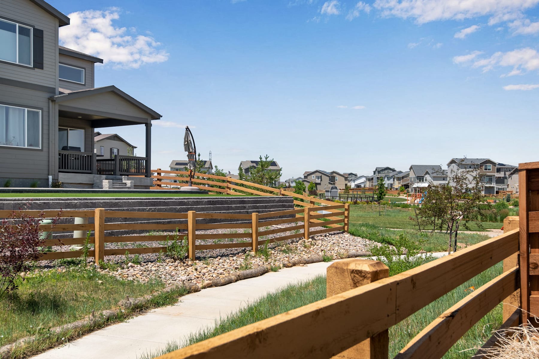 A residential neighborhood with wooden fences, a grassy area, and houses in the background under a cloudy blue sky.