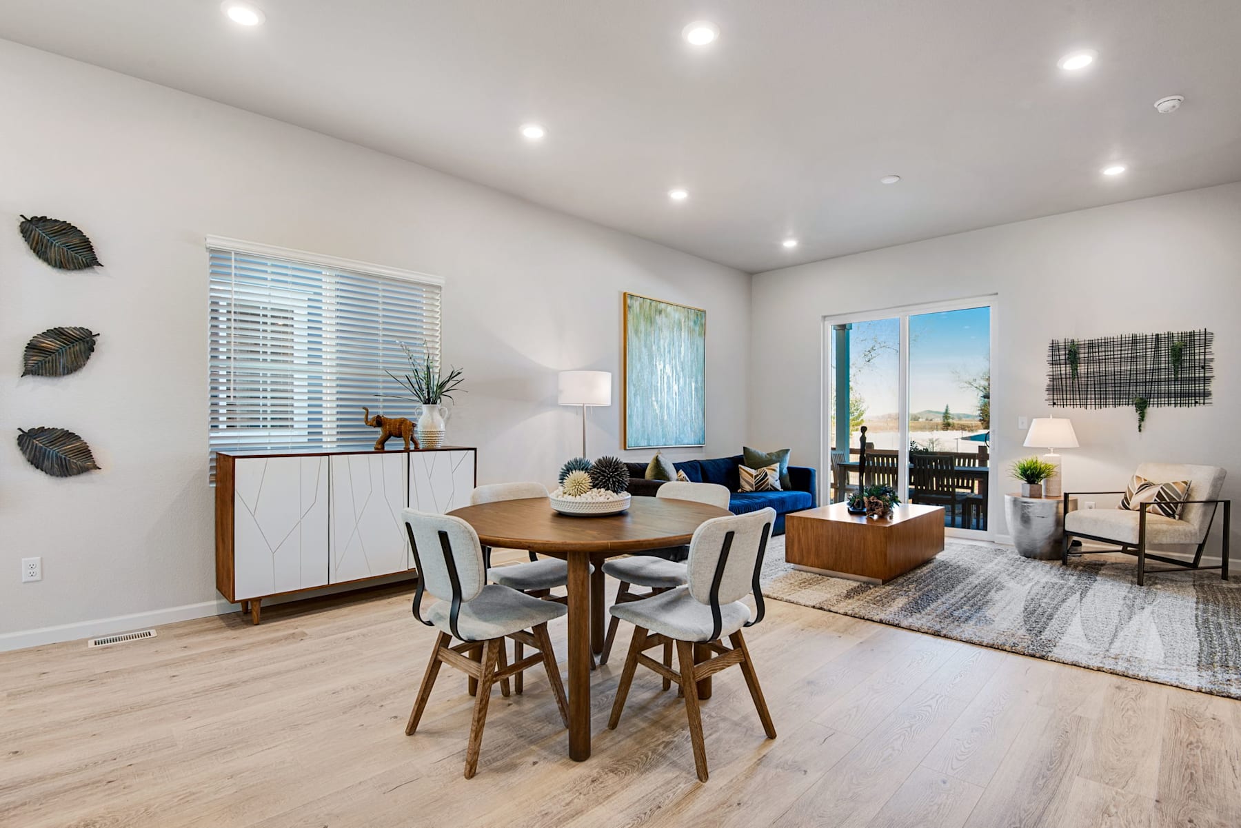 A modern and minimalist dining area with a wooden table and chairs, surrounded by a living room with a couch, shelves, and decorative elements, all illuminated by natural light from the window.