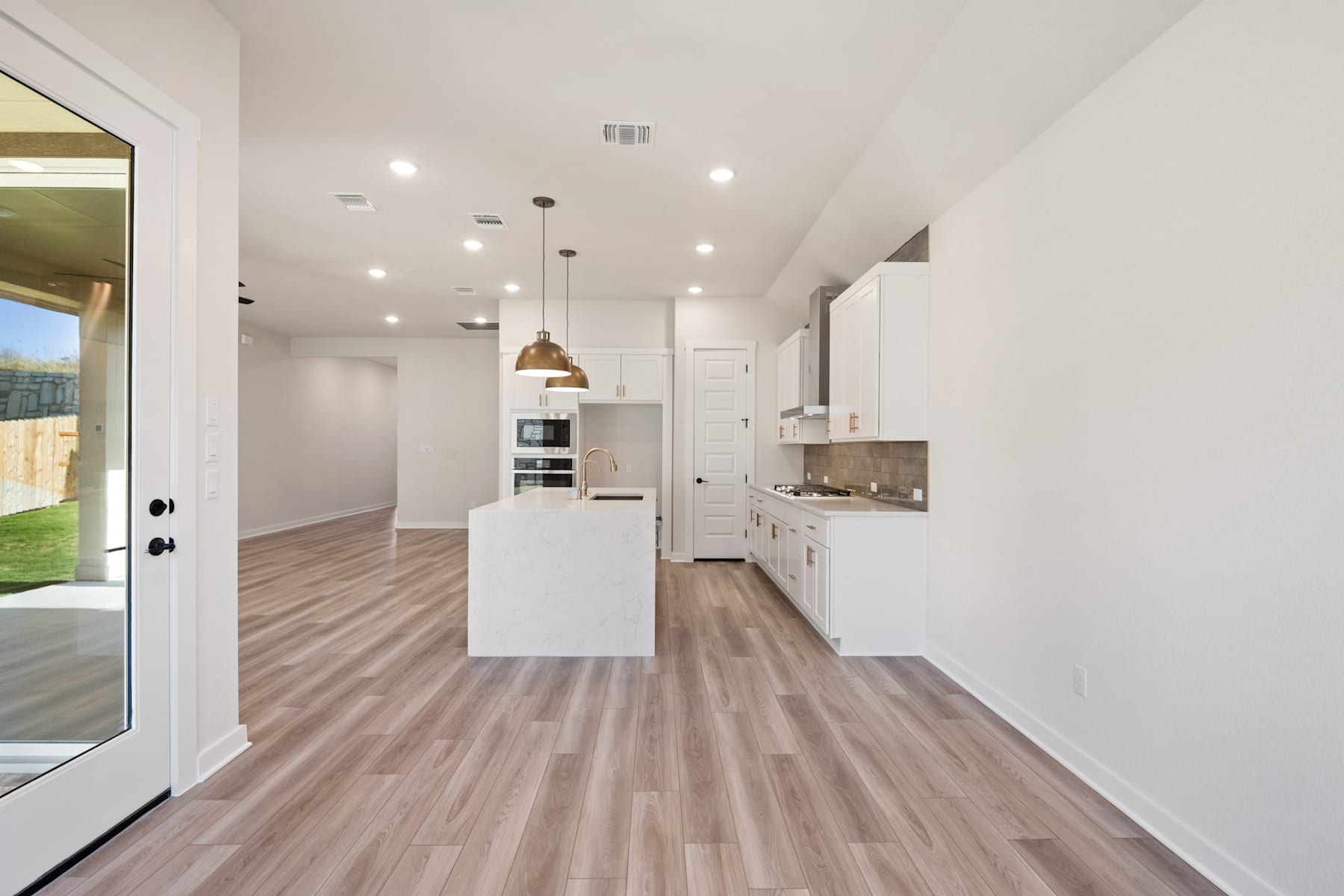 A modern, open-concept kitchen with white cabinets, a central island, and hardwood-style flooring, leading to a bright and airy living space with a sliding glass door to the exterior.
